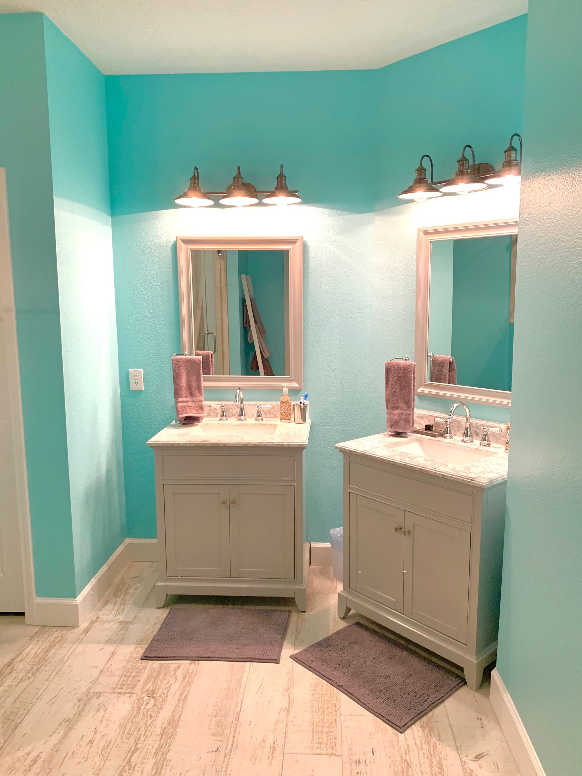 Turquoise bathroom with two white vanities, large mirrors, and wall sconces on either side.