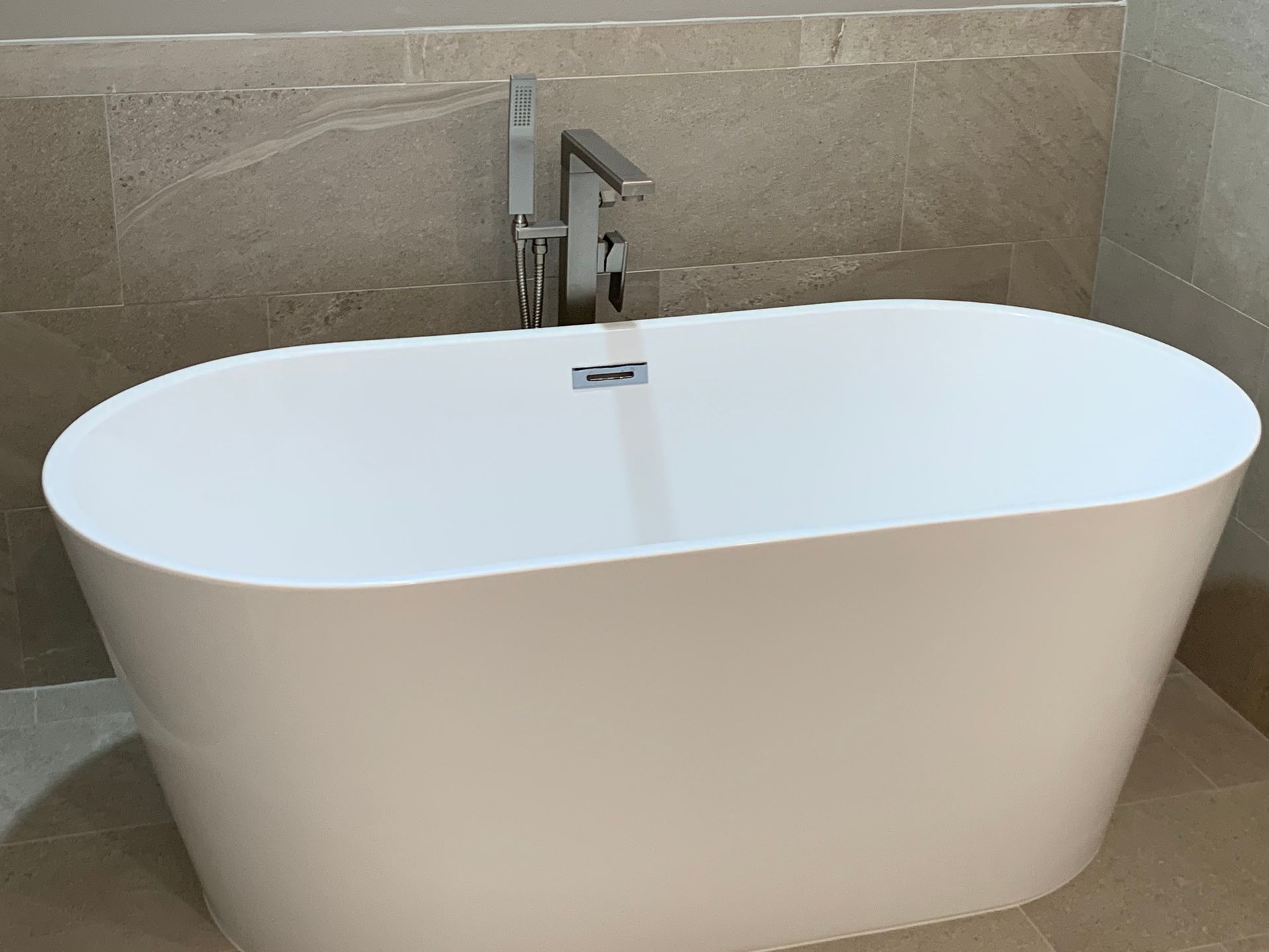 Modern white freestanding bathtub against beige tiled bathroom wall, with chrome faucet.