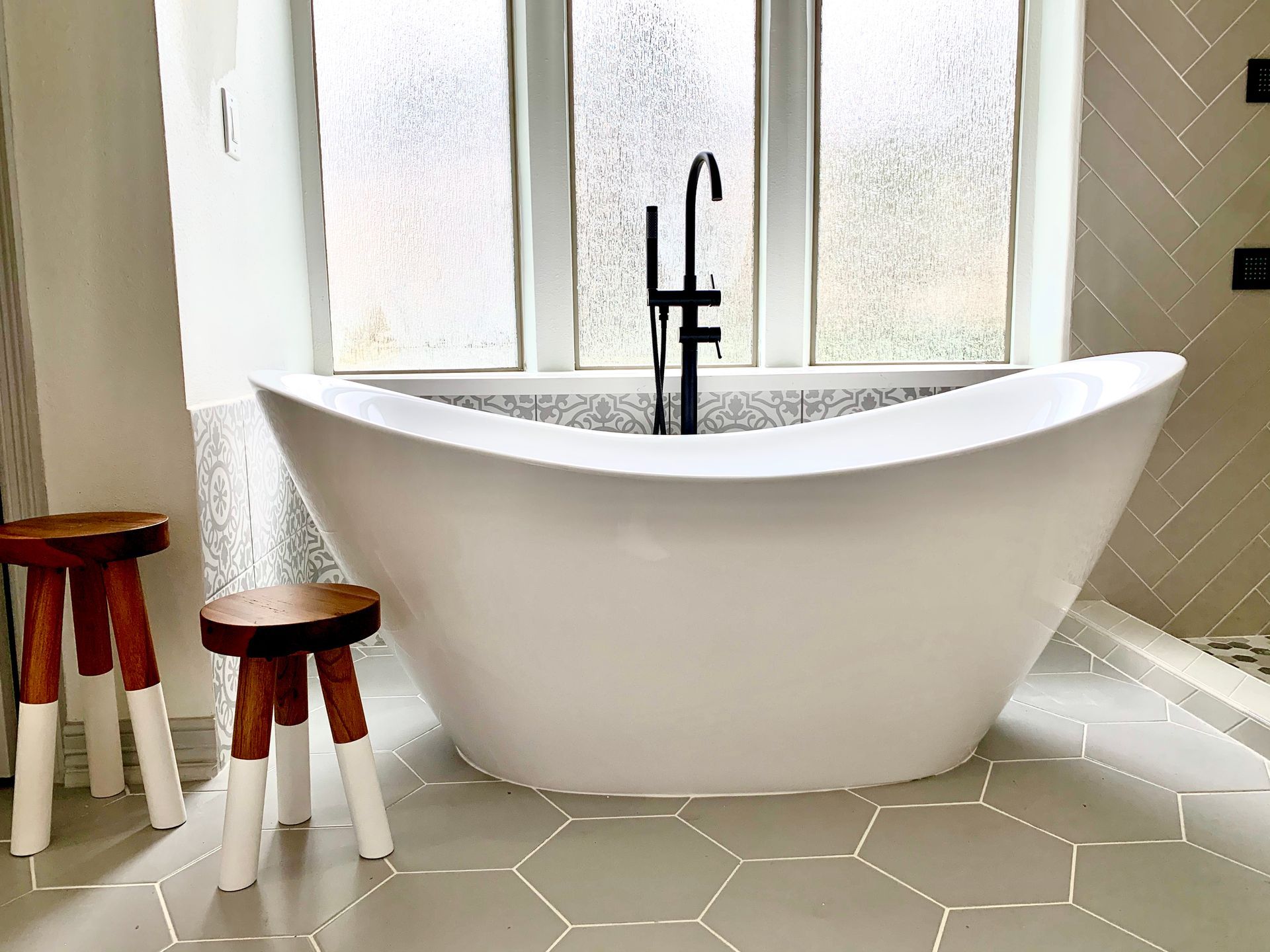 Modern bathroom with a freestanding white tub, black faucet, frosted windows, and two wooden stools