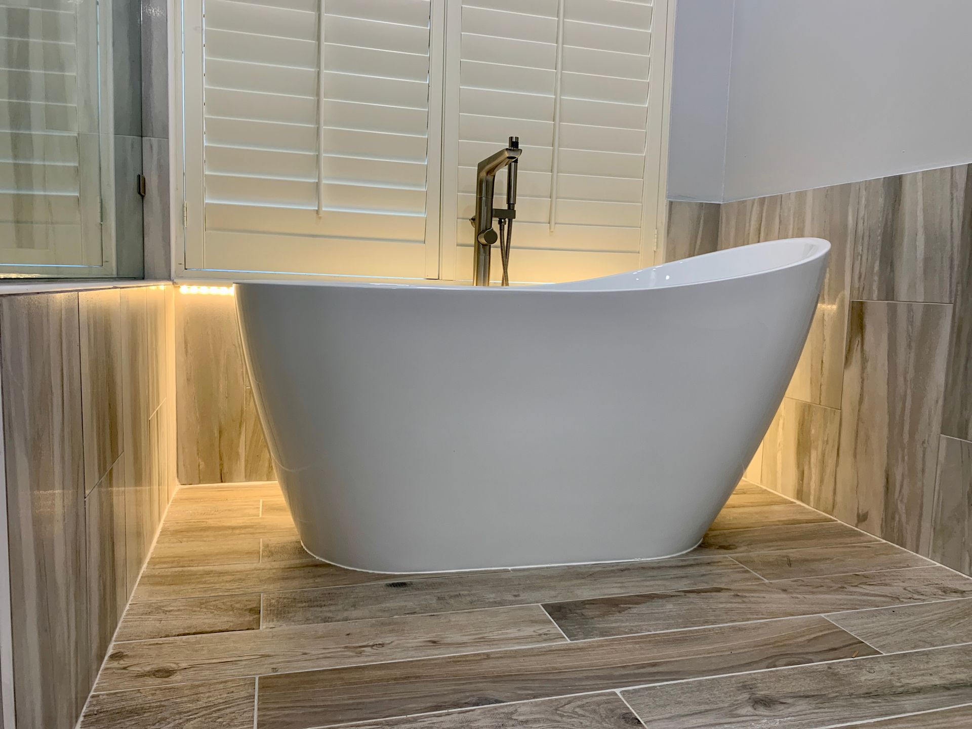 Freestanding white bathtub in a tiled bathroom with shutters and warm underlighting.