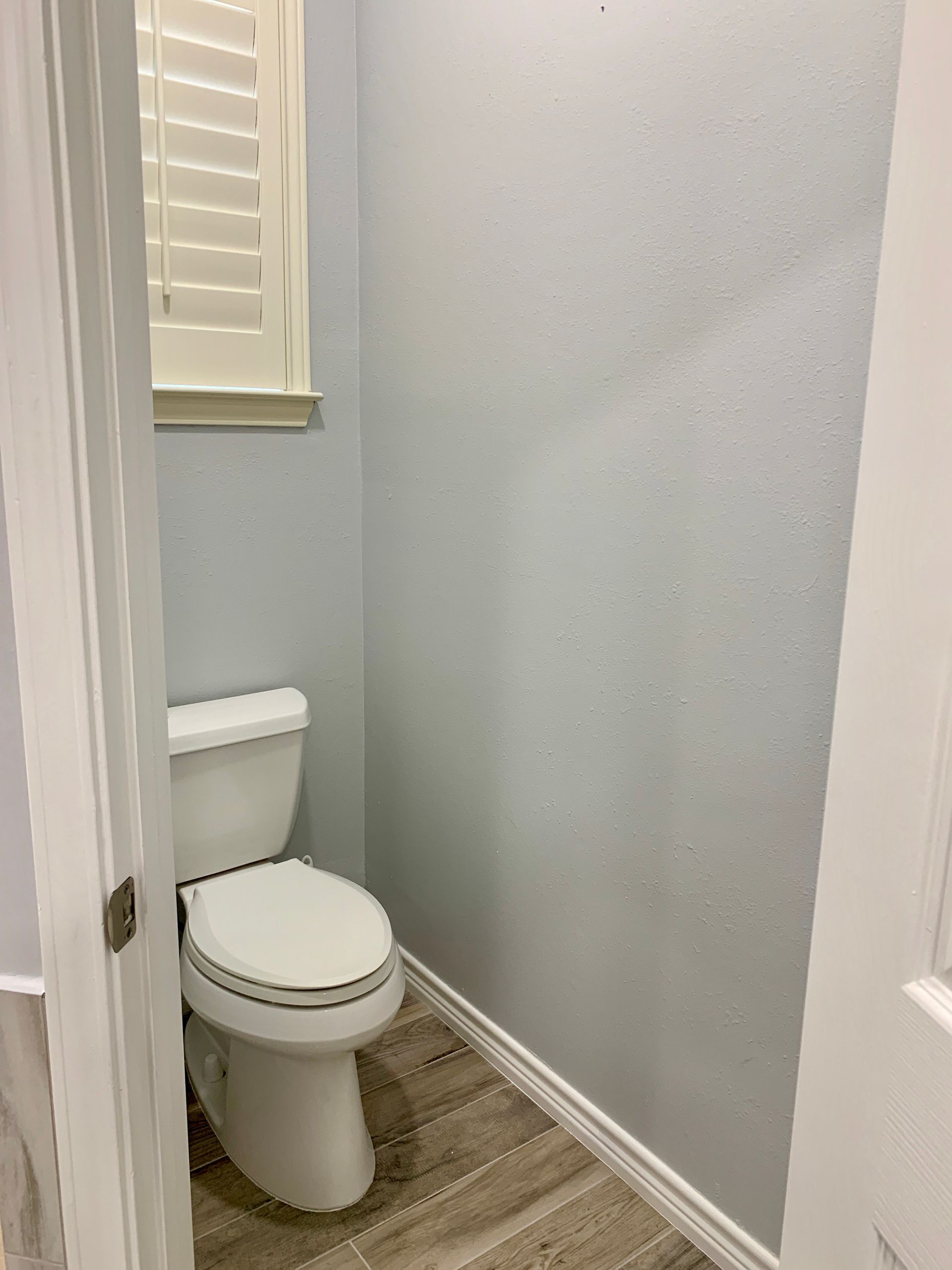 Small bathroom corner with a white toilet, gray walls, wood-pattern floor, and a window with blinds.