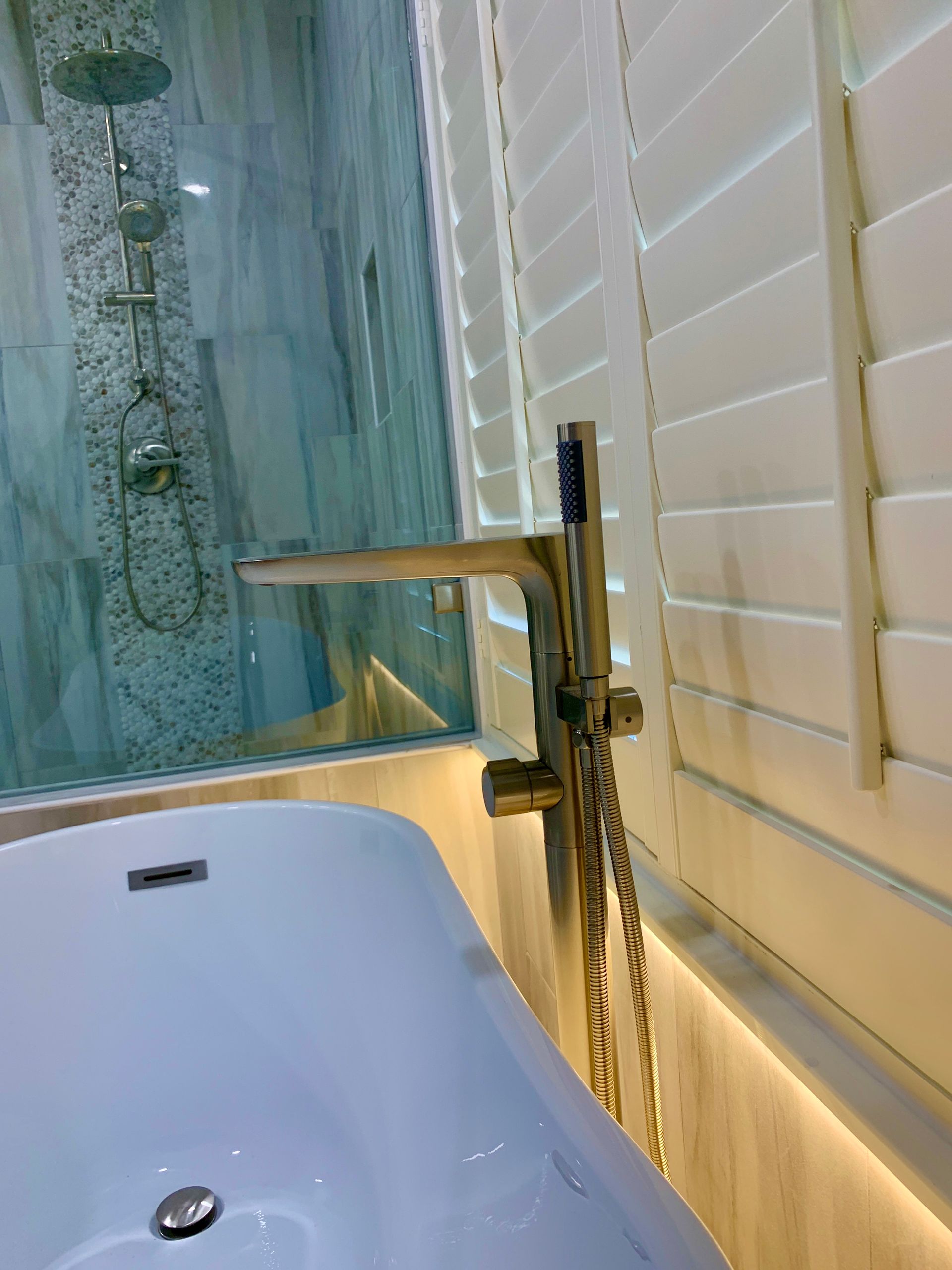 Bathroom with white bathtub, glass shower, chrome faucet, and white shutters beside warm lighting