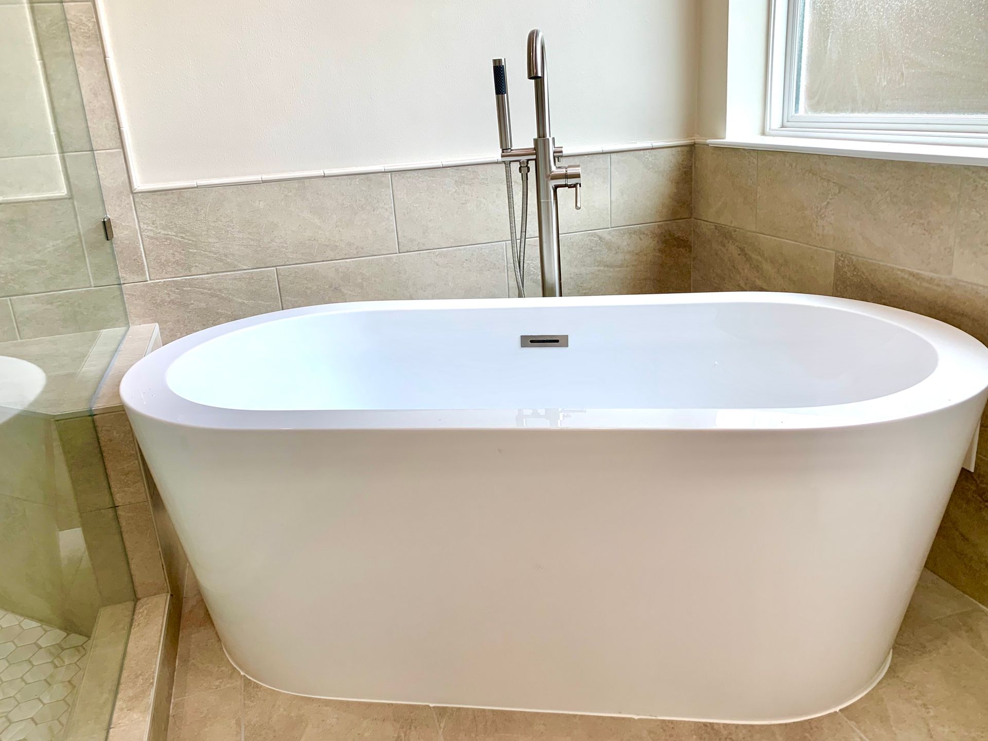 Modern white freestanding bathtub in a tiled bathroom with chrome floor-mounted faucet