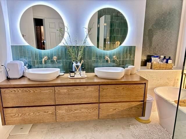 Modern bathroom with double wooden vanity, round mirrors, white sinks, and a freestanding tub by a teal tiled wall