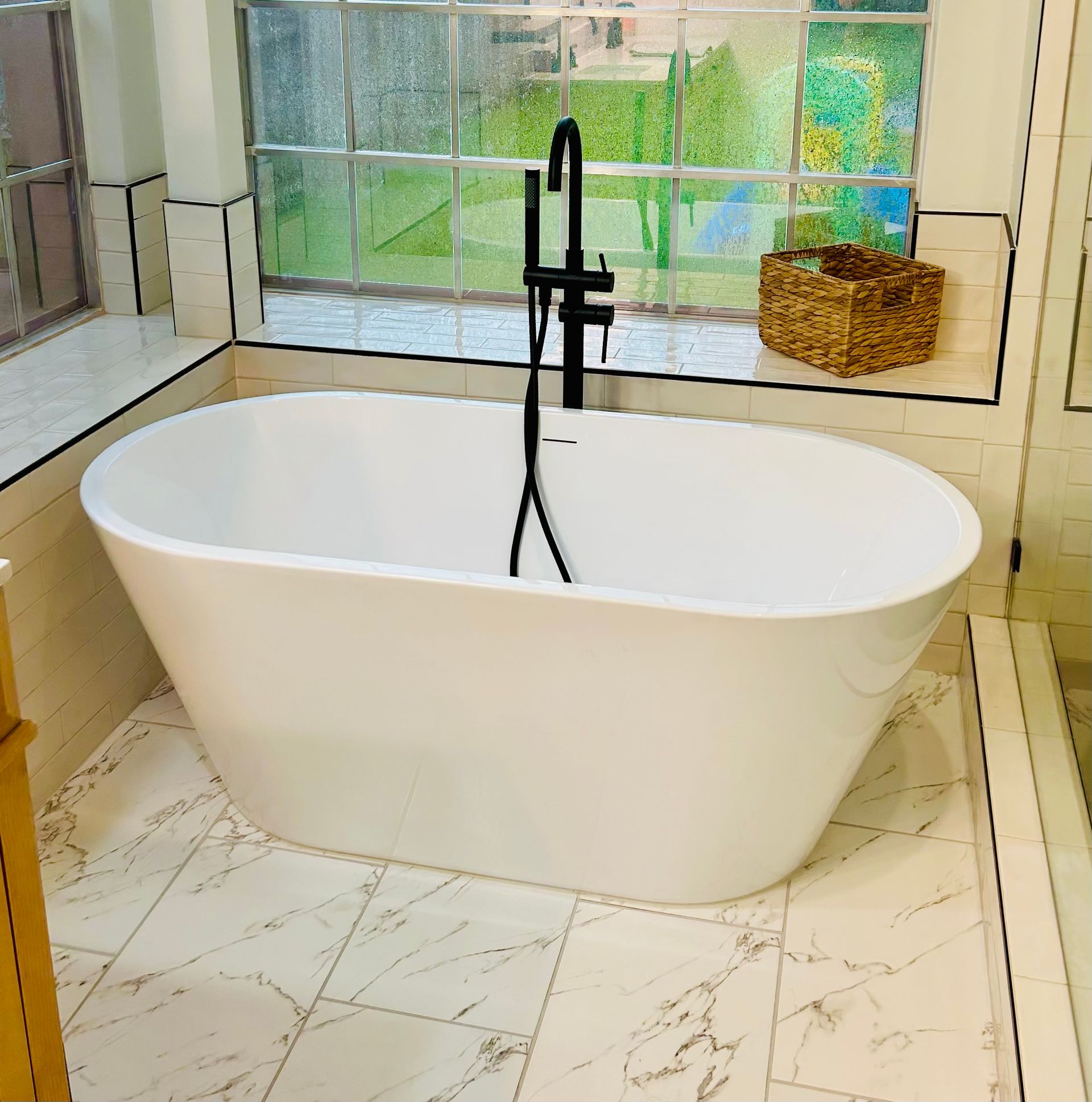 White freestanding bathtub in a bright tiled bathroom by a window and black floor-mounted faucet