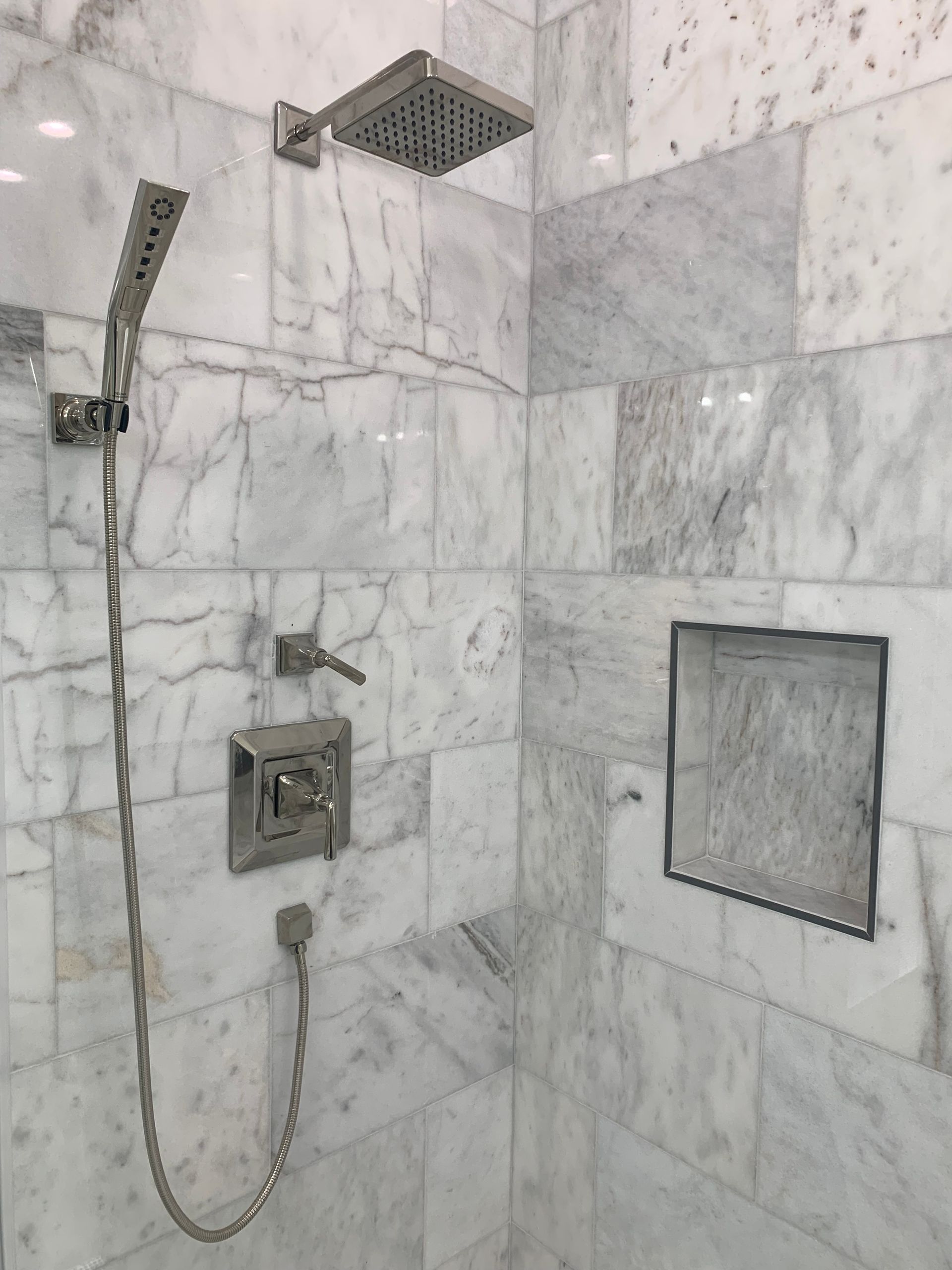 Marble-tiled walk-in shower with square rain showerhead, handheld sprayer, and recessed shelf.