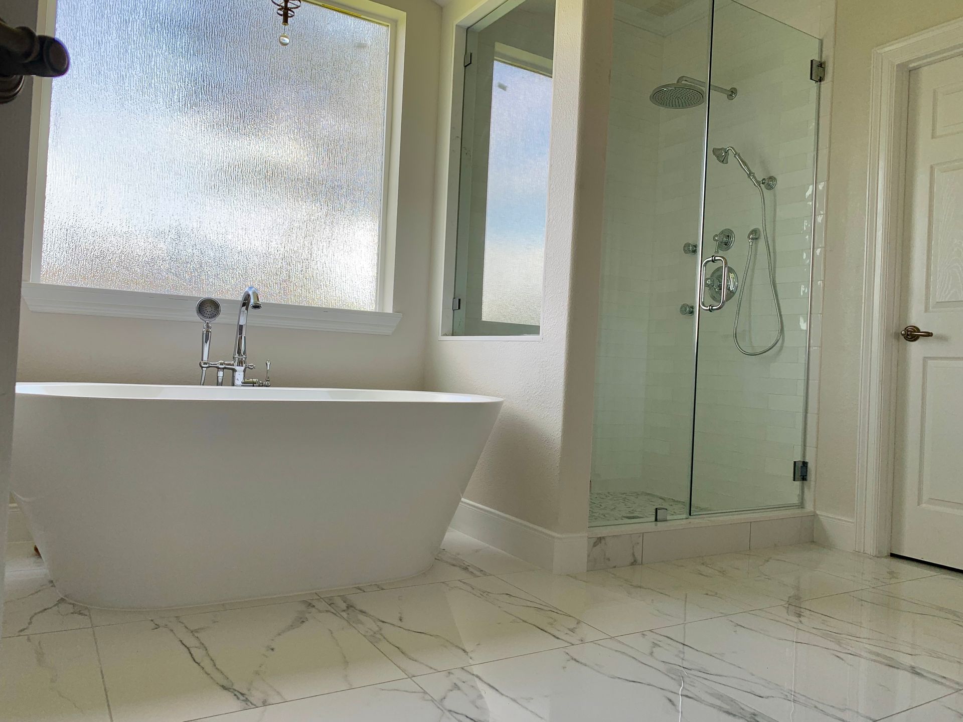 Bright modern bathroom with a freestanding tub, frosted window, glass shower, and white tiled floor.