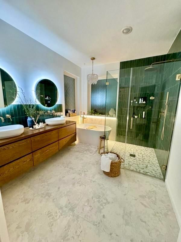 Modern bathroom with wood vanity, illuminated round mirrors, freestanding tub, and glass shower with green tile accent.