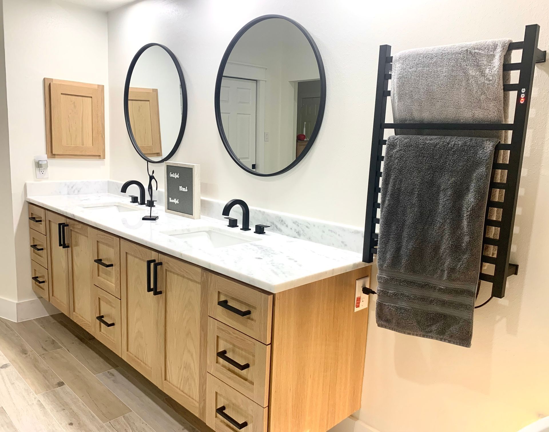 Modern double-sink bathroom vanity with wood cabinets, oval mirrors, and black towel rack with gray towel