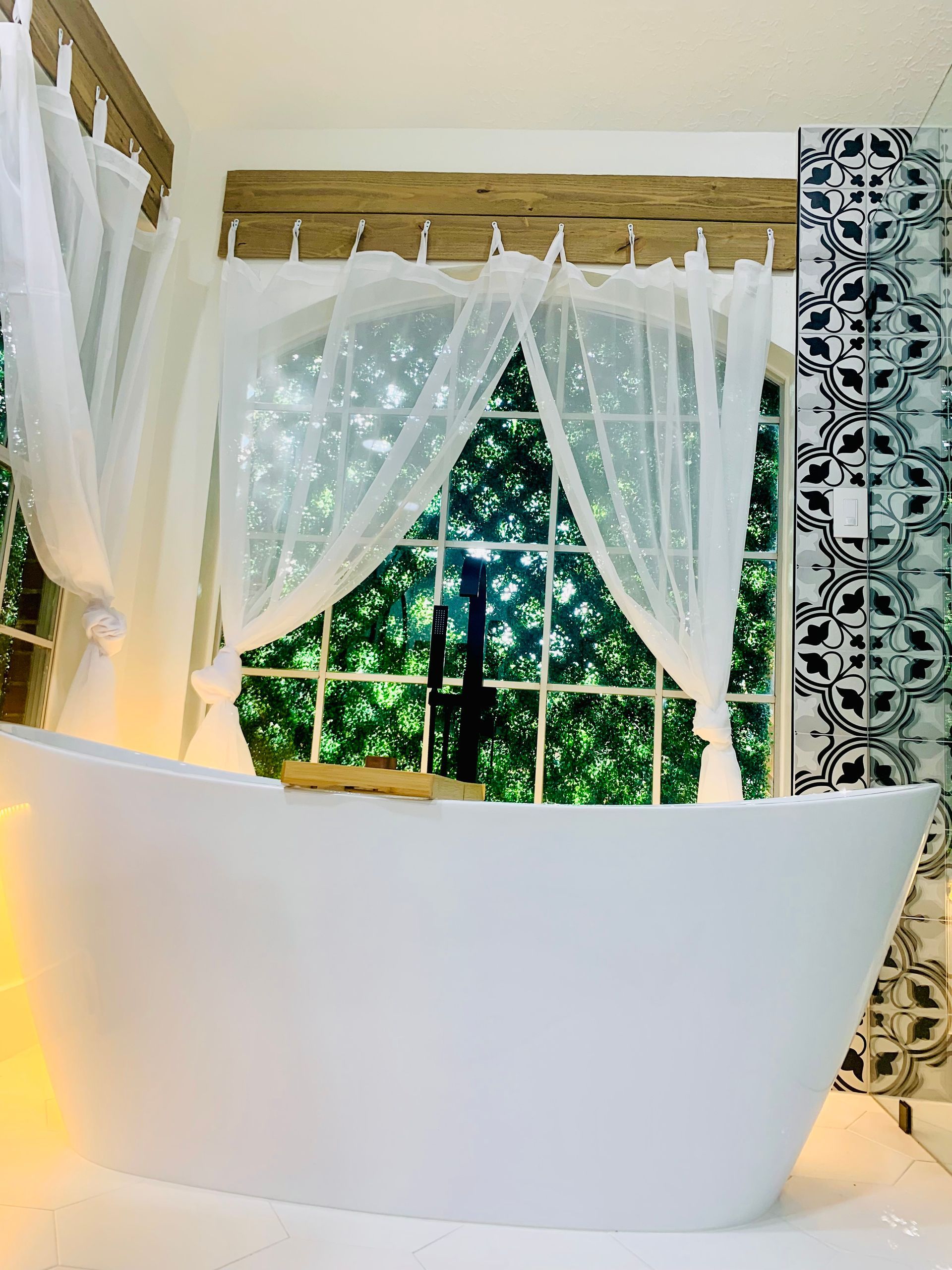 Elegant white freestanding bathtub beneath a window with sheer curtains and leafy greenery outside
