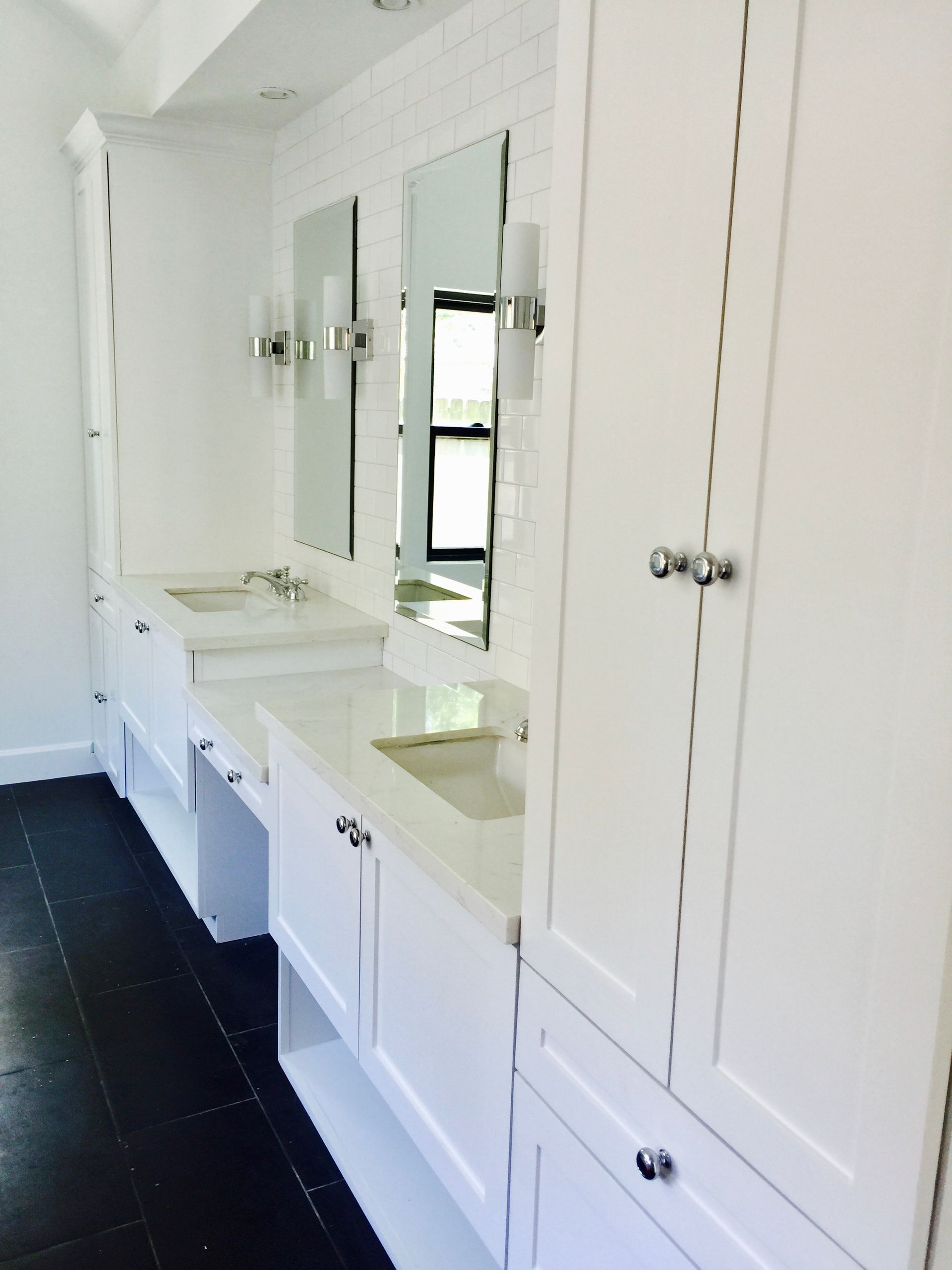 White bathroom vanity with mirrors, cabinets, and dark floor tiles against a bright wall.