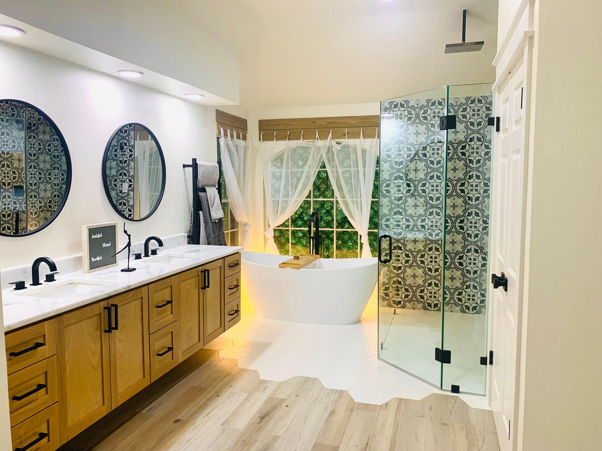 Modern bathroom with double vanity, freestanding tub by a window, and glass shower with patterned tile.