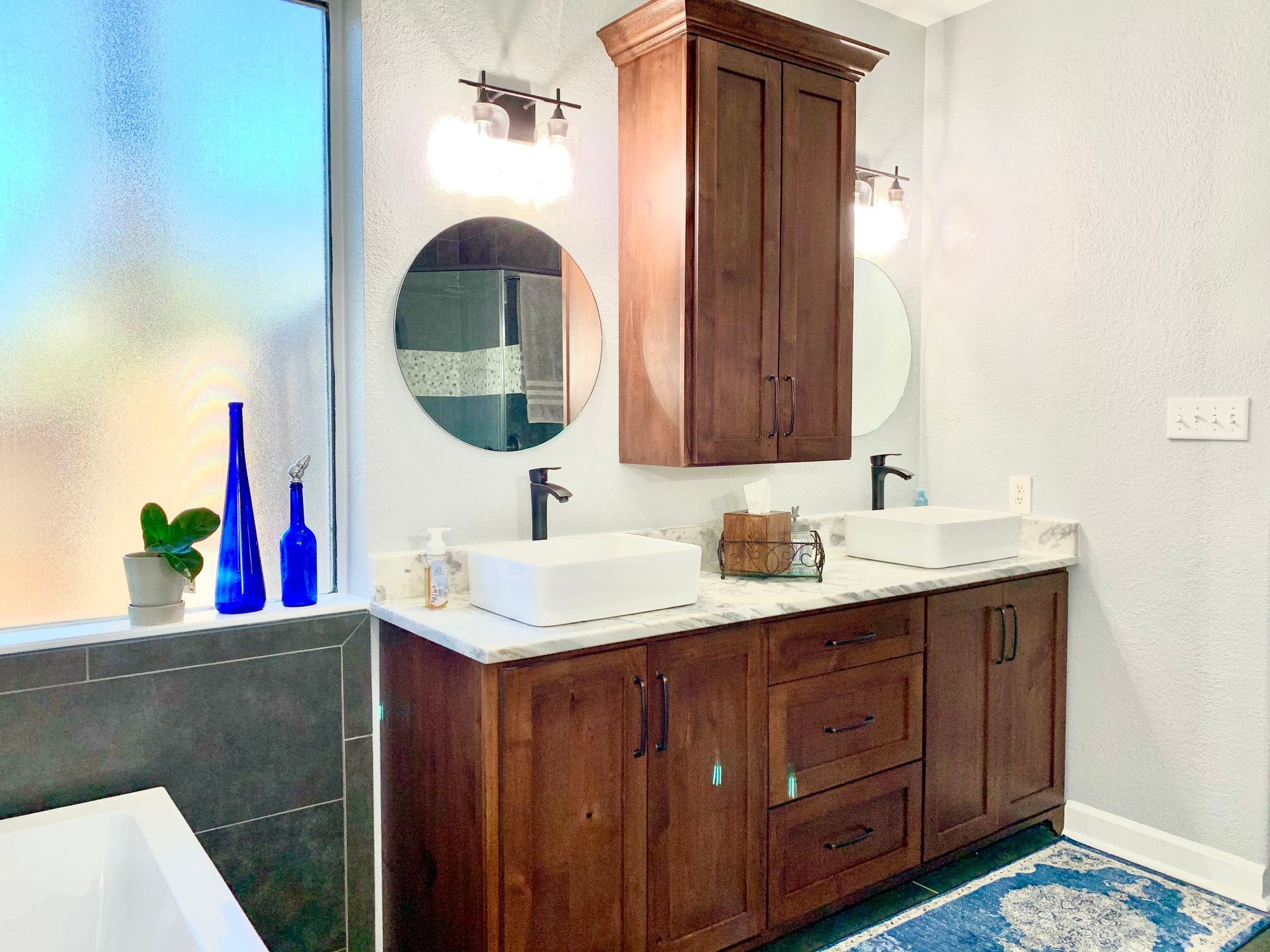 Bathroom vanity with dark wood cabinets, white sink, round mirror, and blue glass vases on the counter