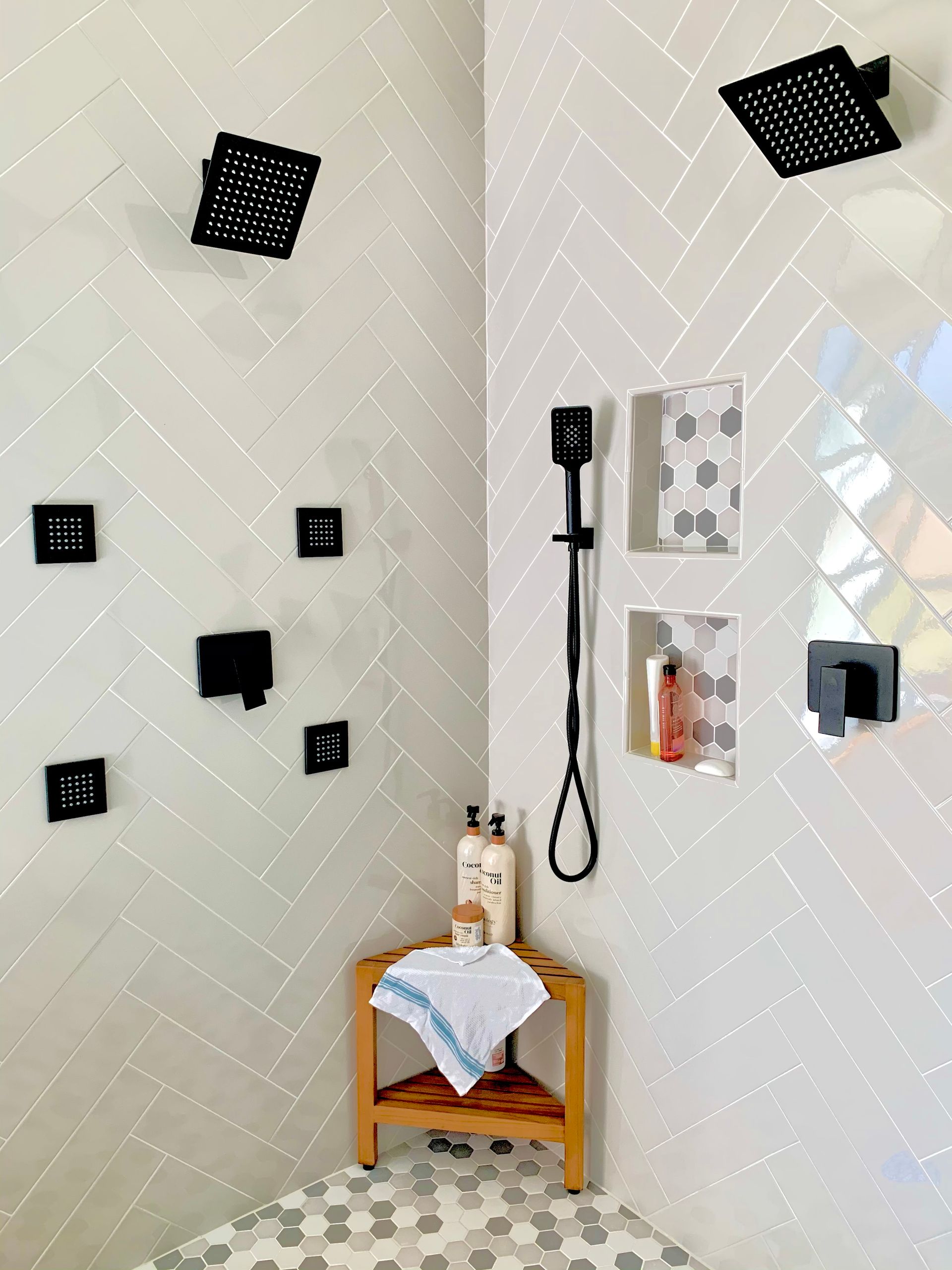 Bathroom corner with shower fixtures, wall-mounted accessories, and a small wooden stool with folded towel and bottles