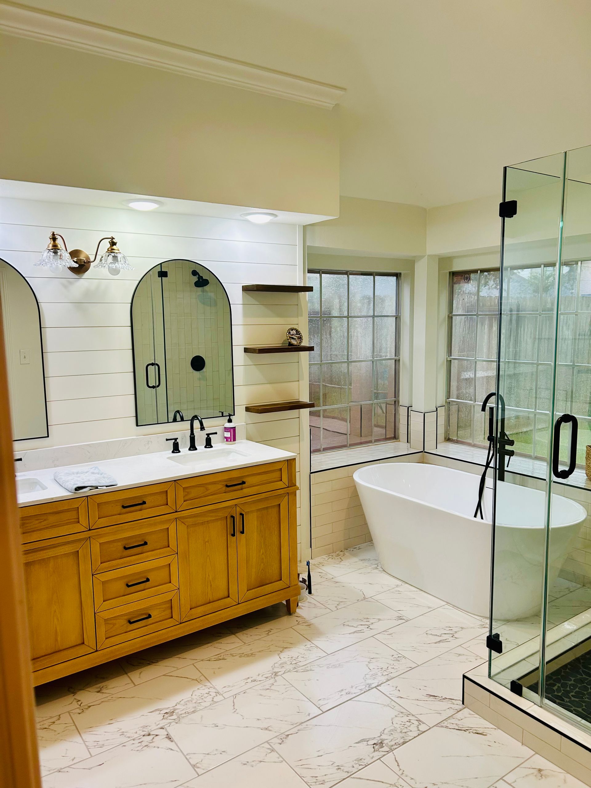 Bright bathroom with wooden vanity, oval mirror, freestanding tub, and glass shower enclosure