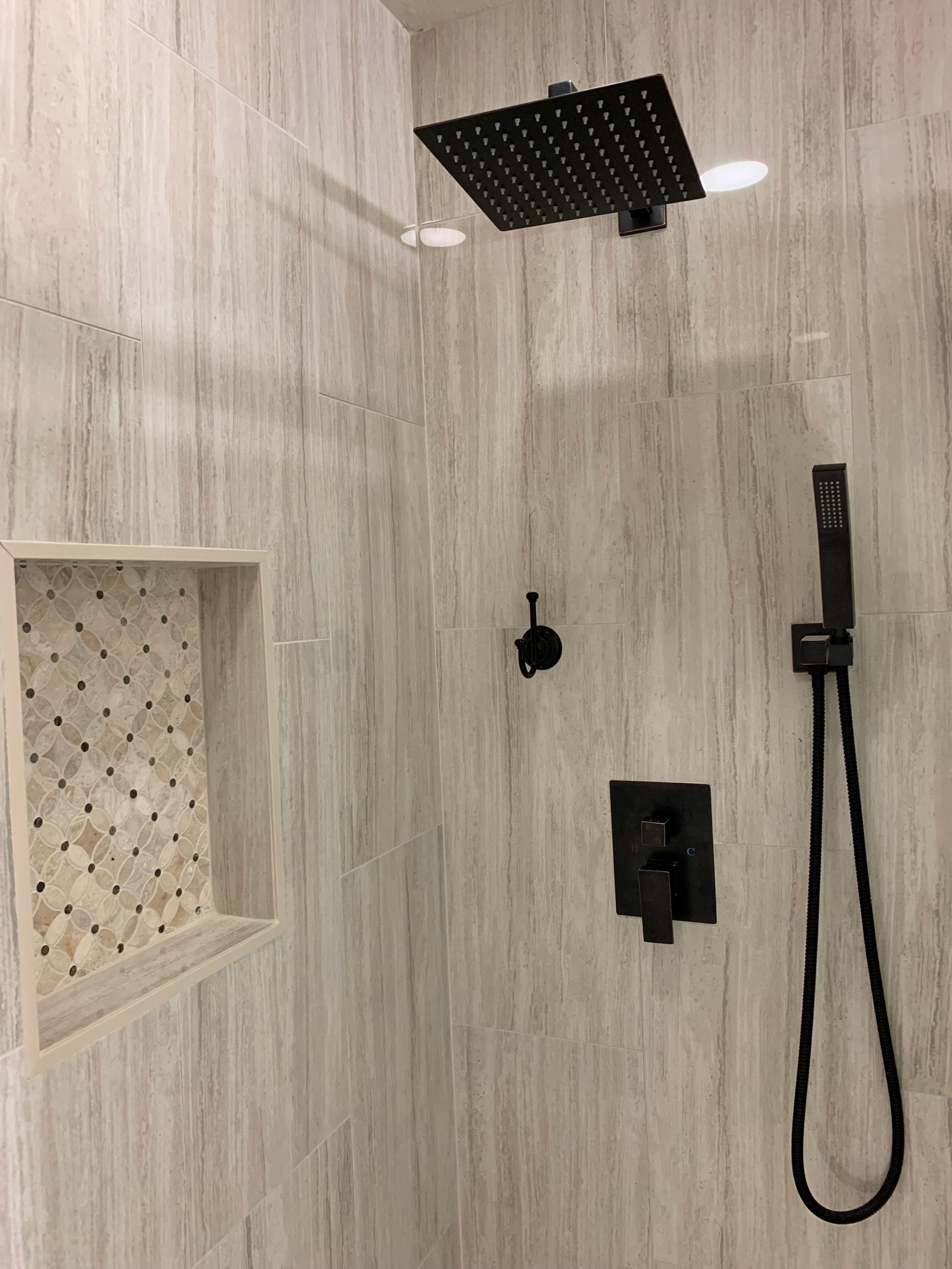 Modern shower with light wood-look tile, black rain showerhead, niche, and handheld sprayer.
