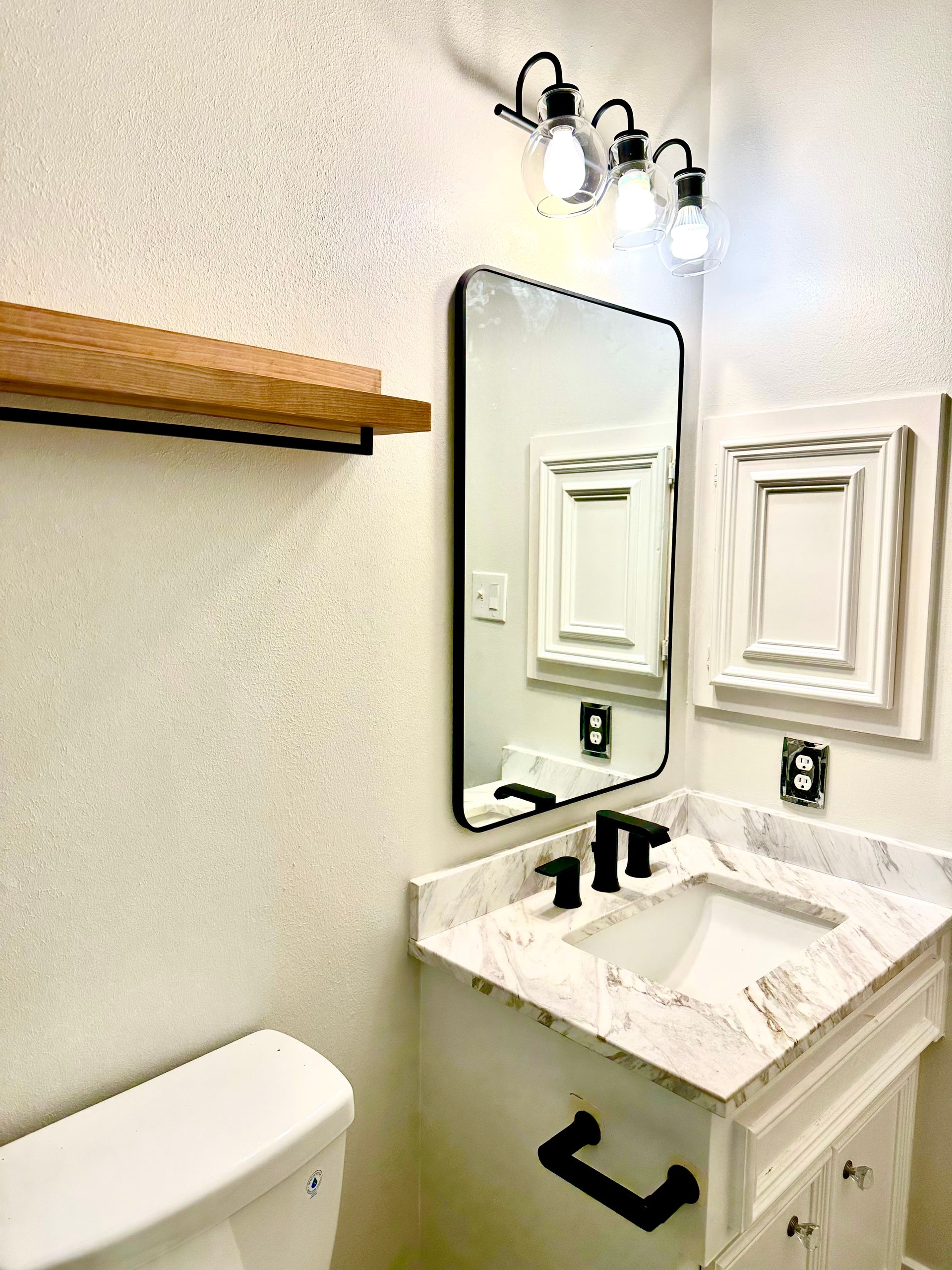 Small white bathroom with a vanity, mirror, wall sconce, towel shelf, and black fixtures.