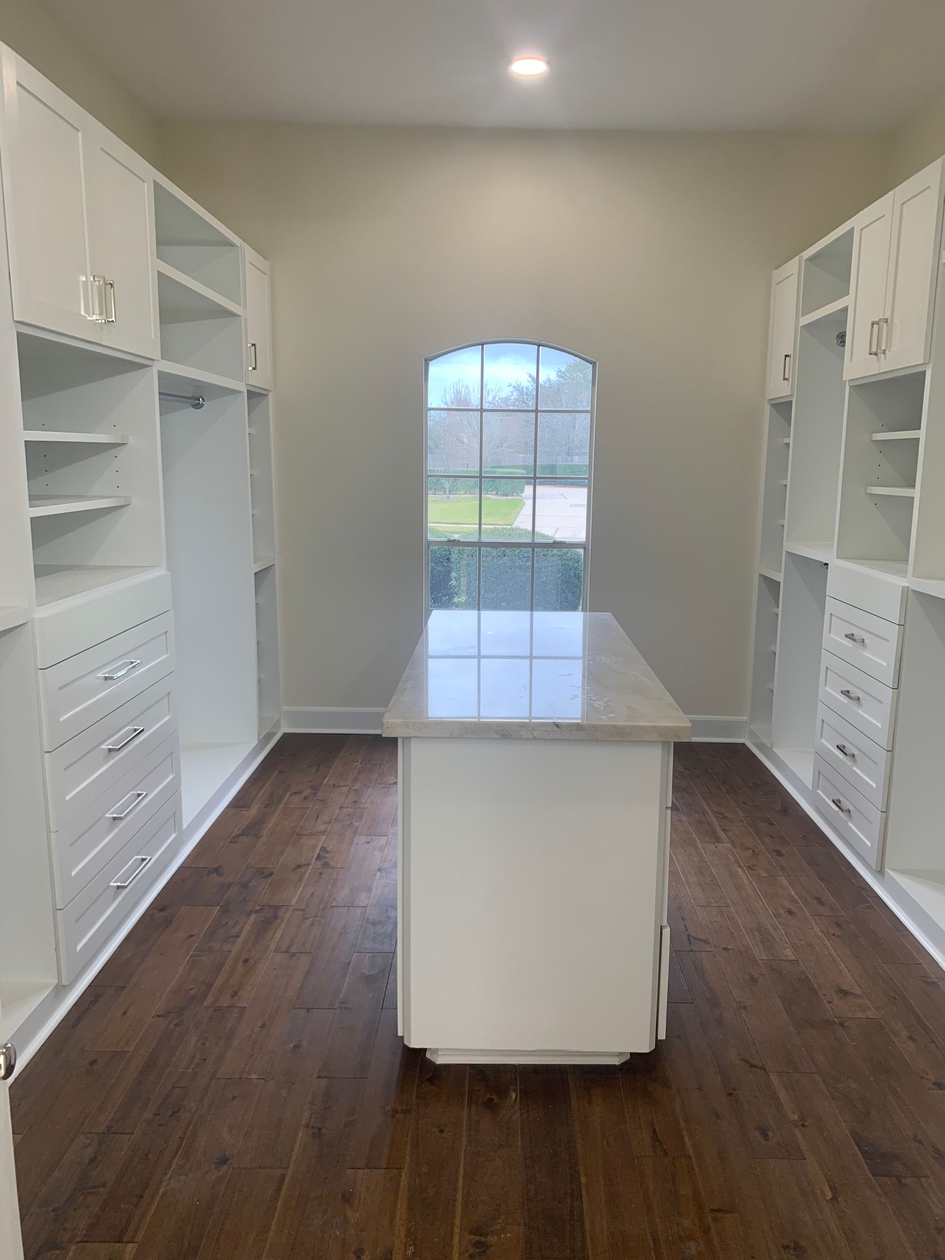 Bright walk-in closet with white built-in shelving, drawers, and an island on dark wood floors.