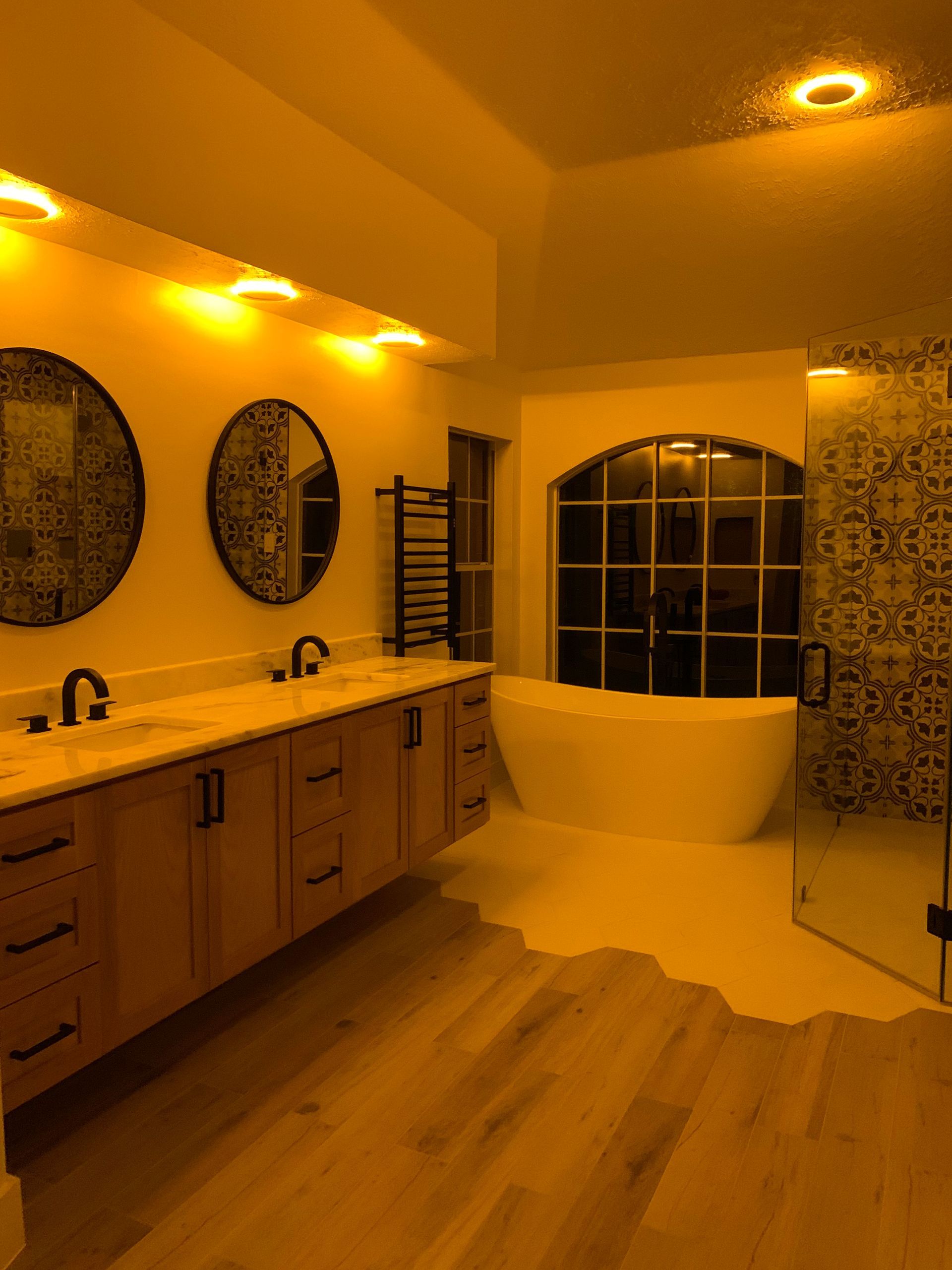 Warm-lit modern bathroom with wood vanity, round mirrors, tub, and patterned shower tiles