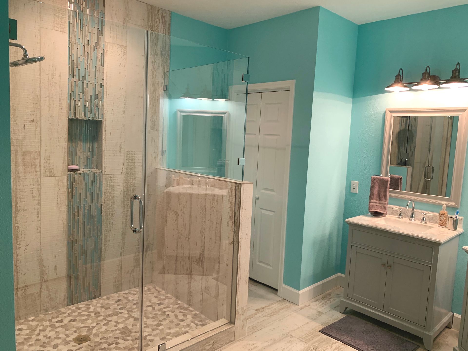 Turquoise bathroom with glass shower, white doors, and a vanity lit by three sconces.