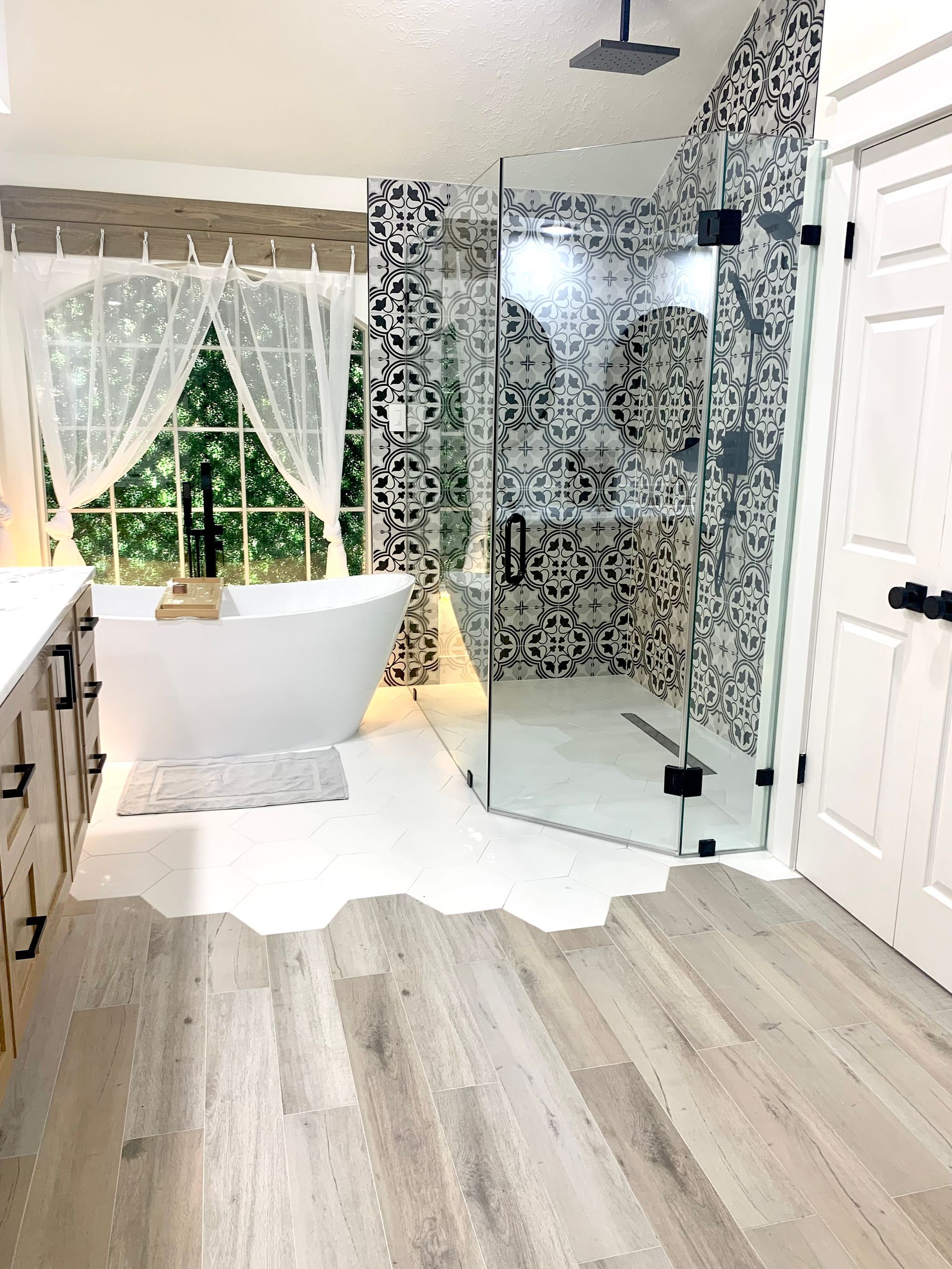 Bright bathroom with freestanding tub, glass shower, patterned tile, and window with white curtains.