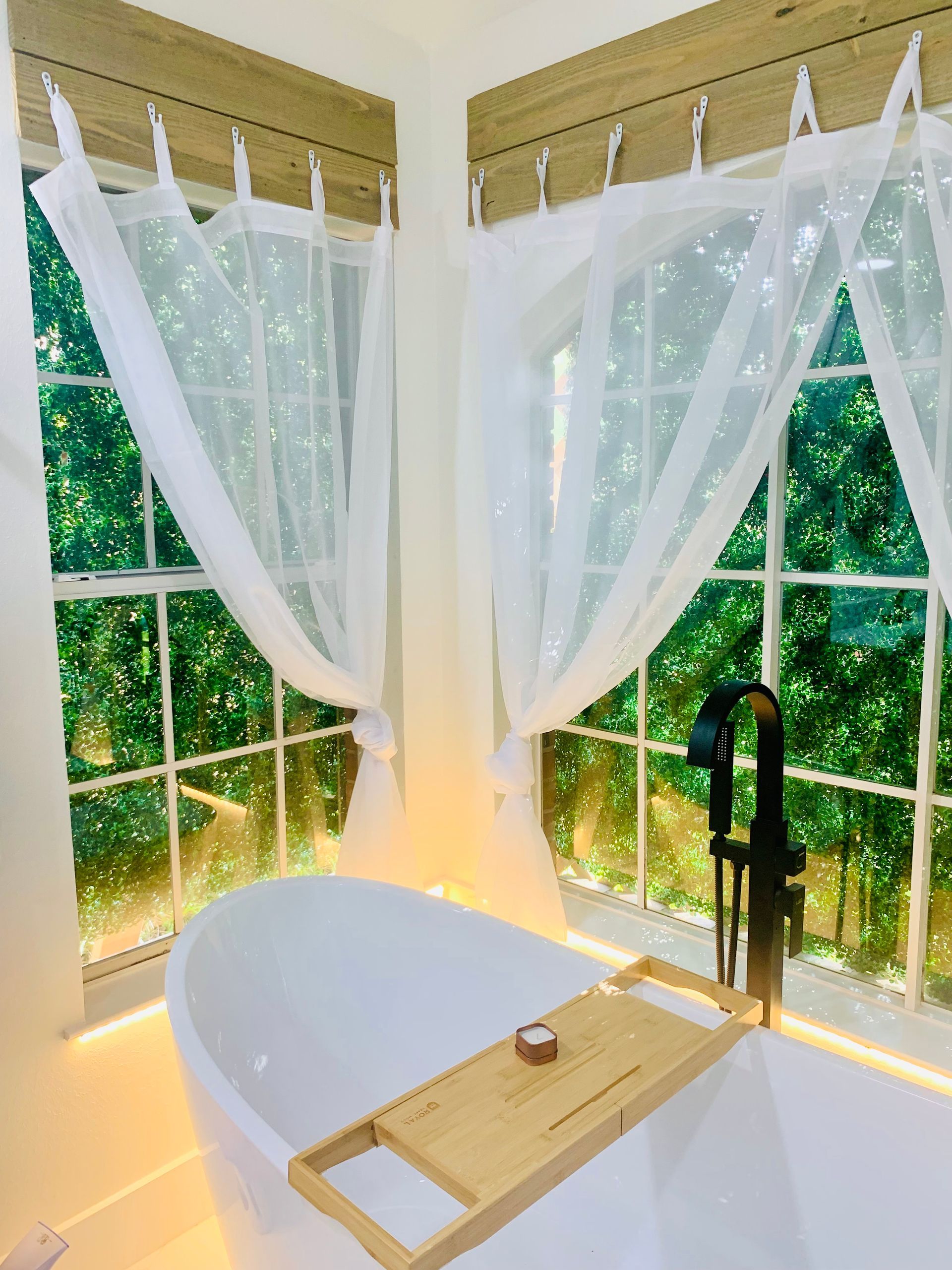 Bright bathroom with a freestanding tub by a window, sheer white curtains, and soft warm lighting