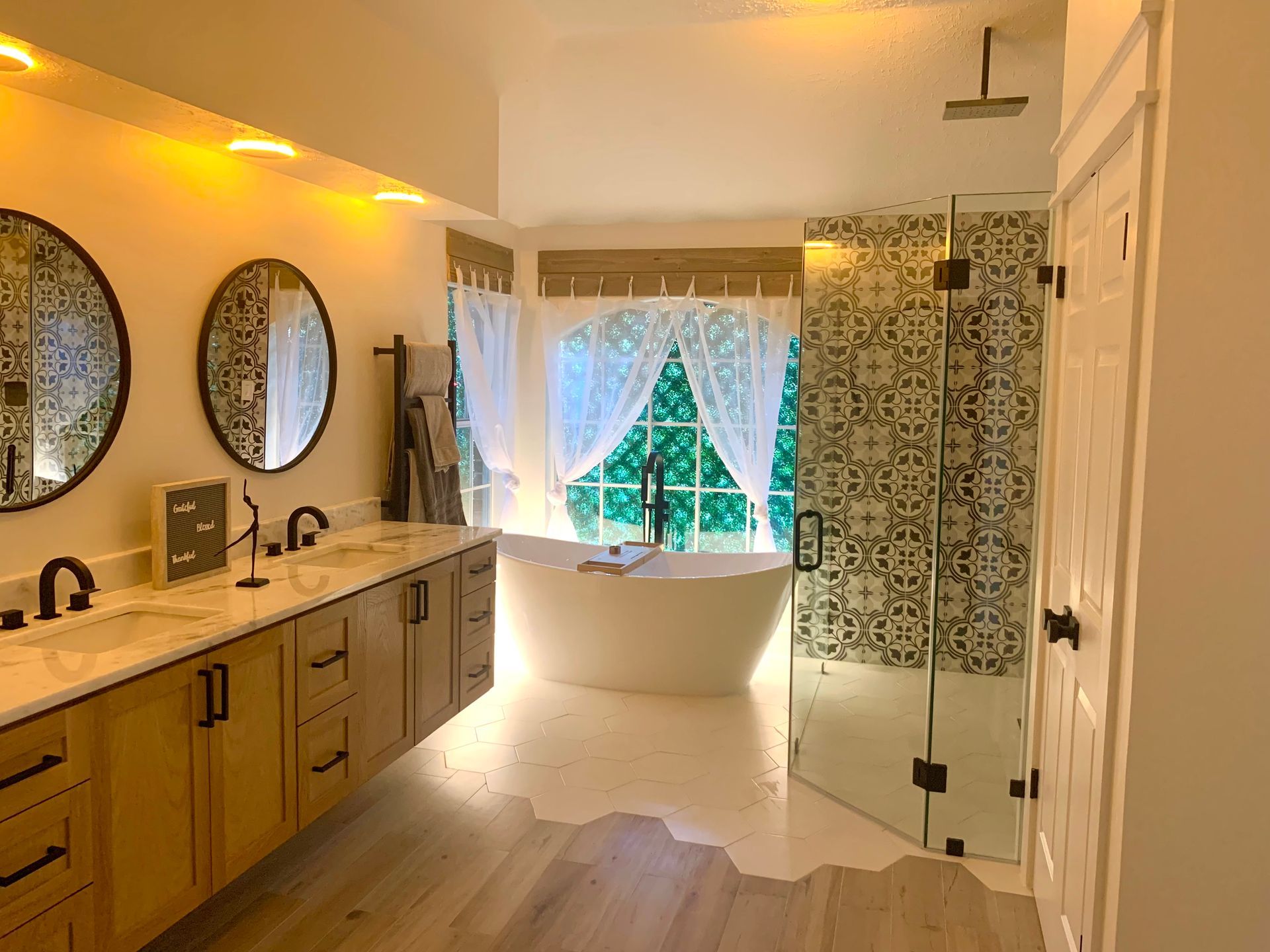 Bright bathroom with twin sinks, circular mirrors, freestanding tub, and patterned shower curtain by a window
