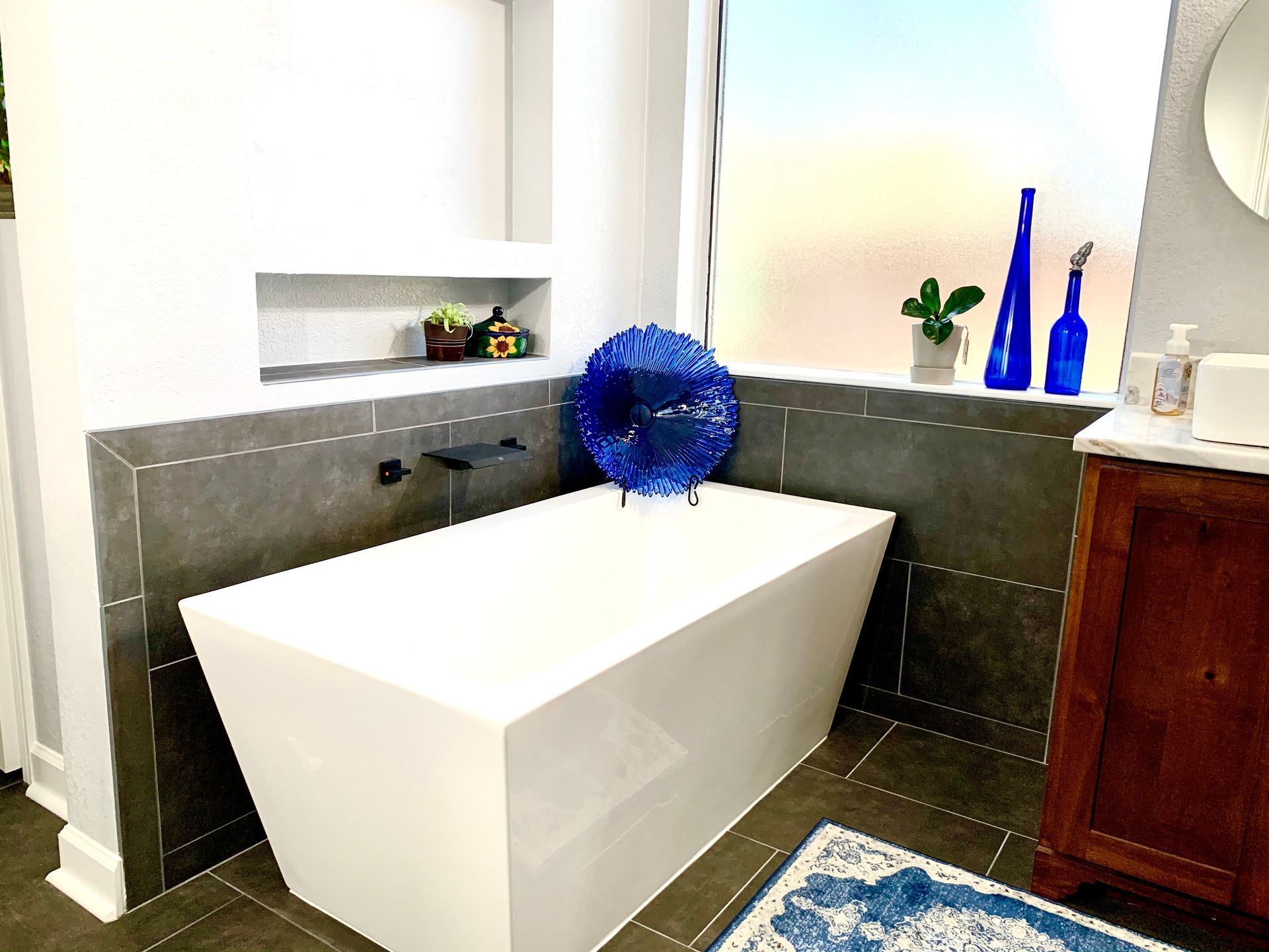 Modern bathroom with a white bathtub, gray tiled surround, and blue decorative bottles on the ledge