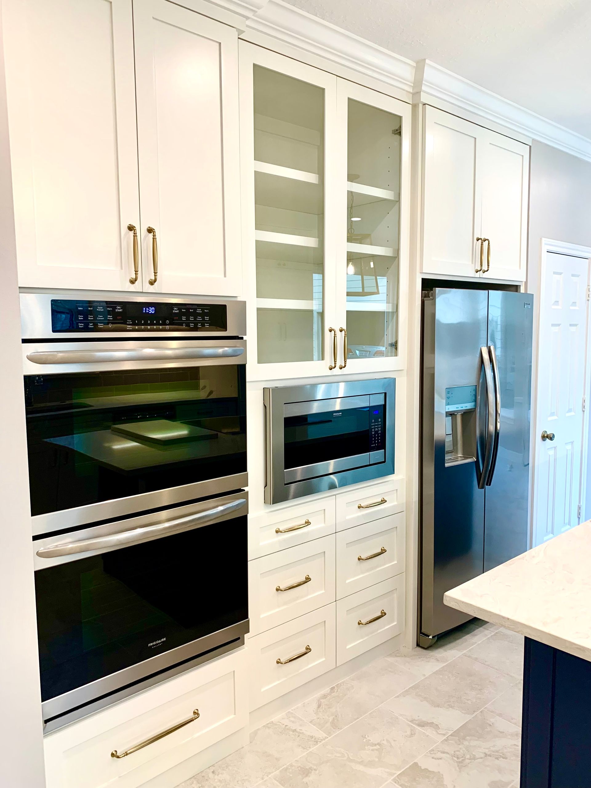 Modern white kitchen with built-in ovens, glass-front cabinets, and a stainless steel refrigerator