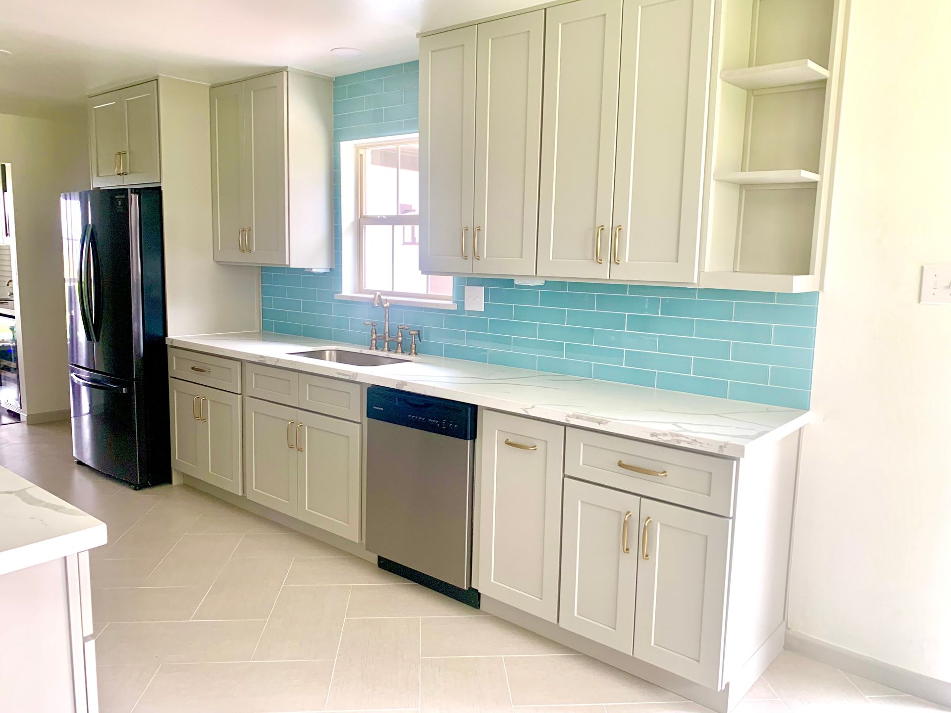 Bright modern kitchen with gray cabinets, blue backsplash, black fridge, and stainless sink and dishwasher