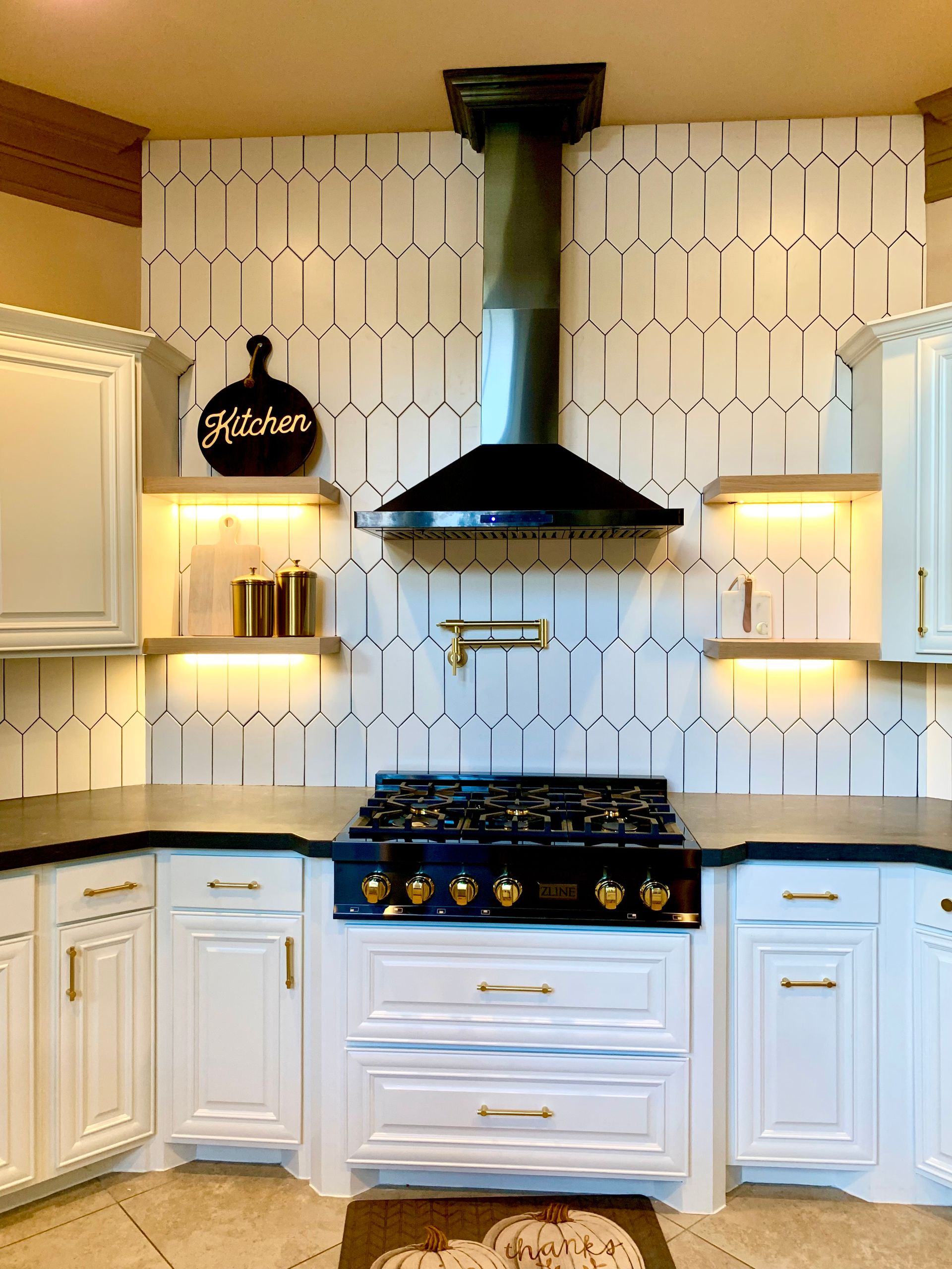 White kitchen with black range hood, patterned tile backsplash, and gold-accented open shelves
