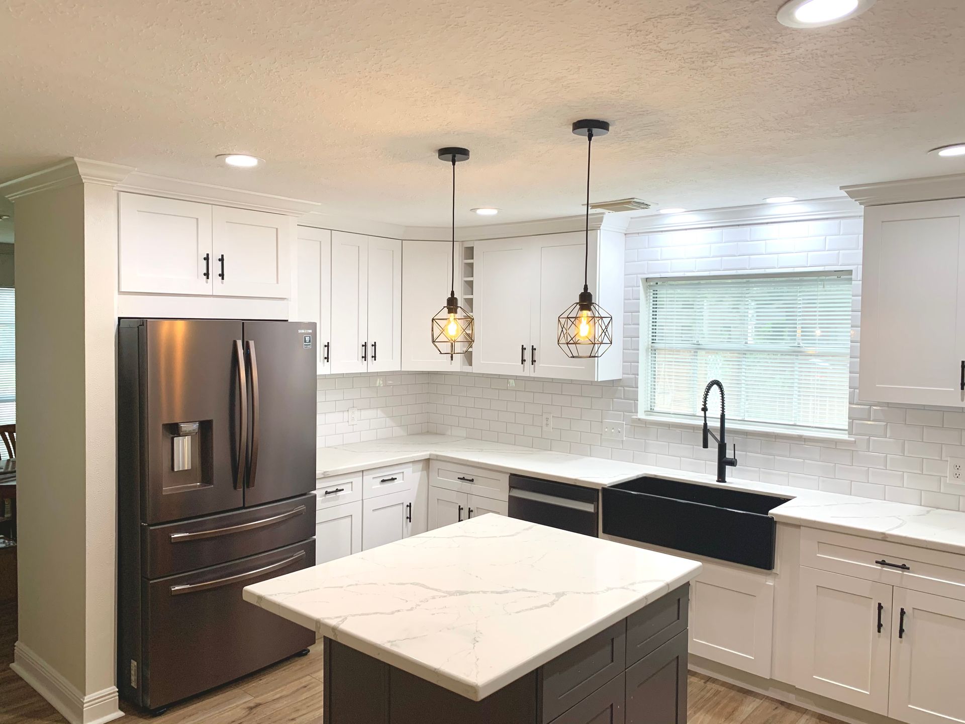 Modern white kitchen with black refrigerator, marble island, pendant lights, and window above sink
