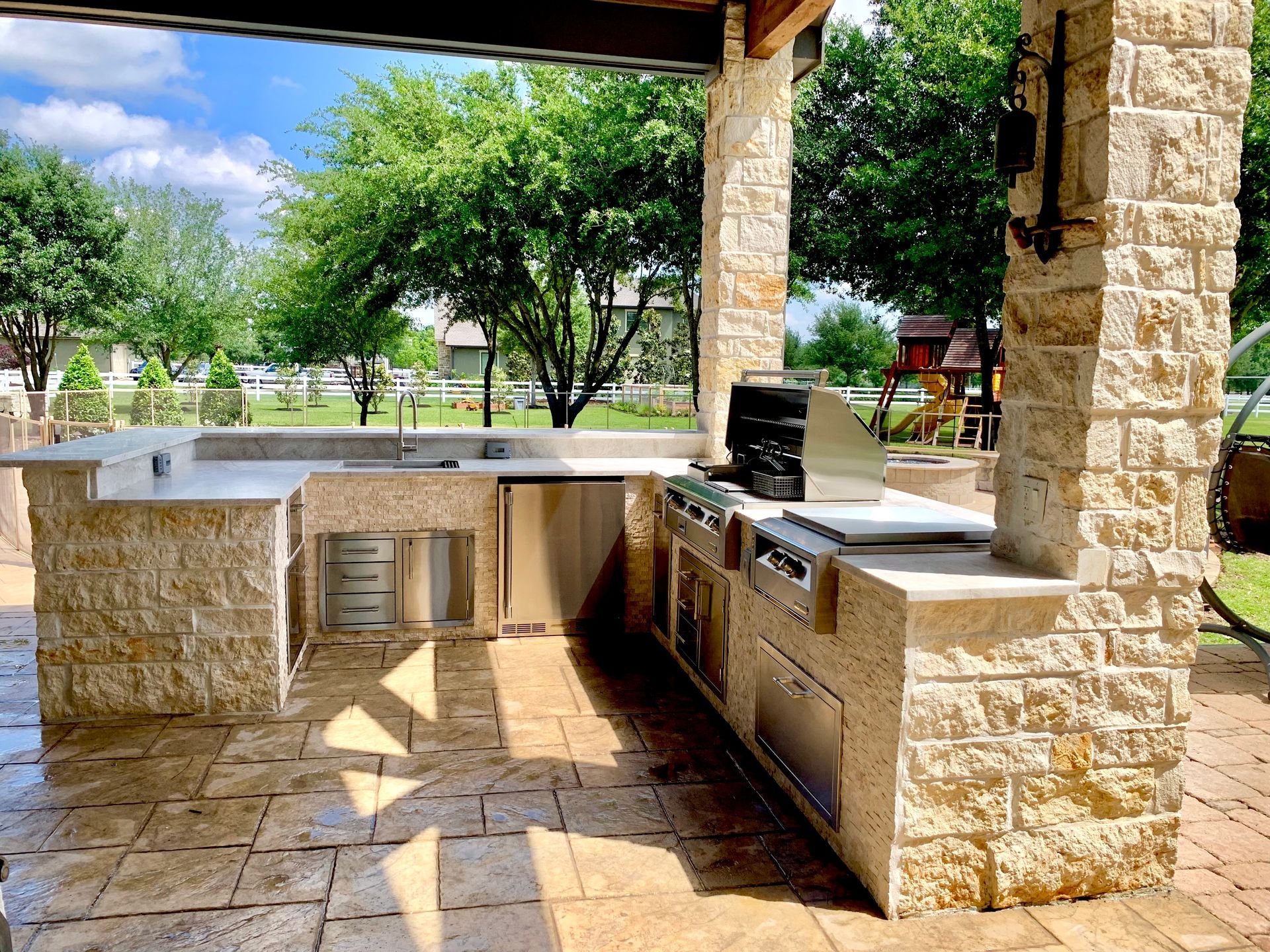 Stone outdoor kitchen with grill and sink under a shaded patio, overlooking a green yard