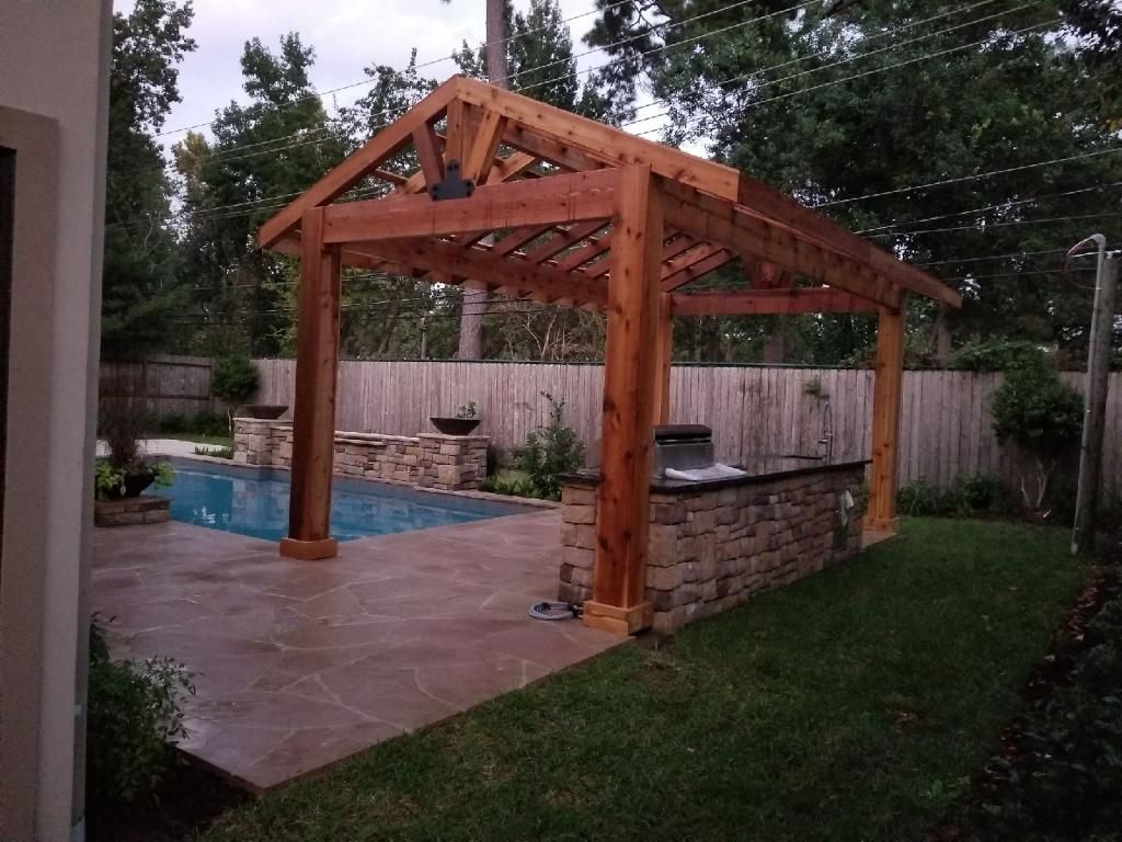 Wooden pergola over a backyard patio beside a small pool and stone wall, surrounded by trees.