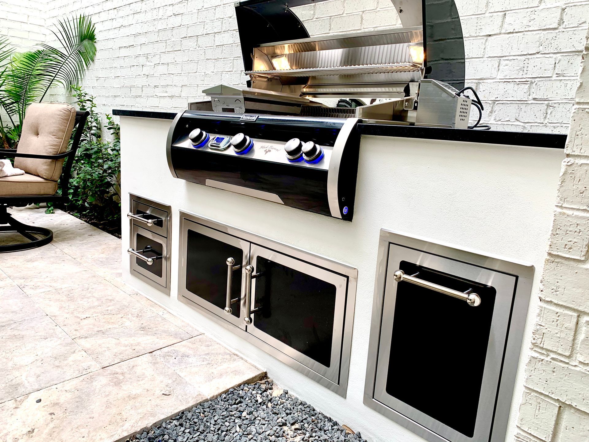Outdoor built-in grill station with stainless steel grill and black cabinet doors against a white brick wall