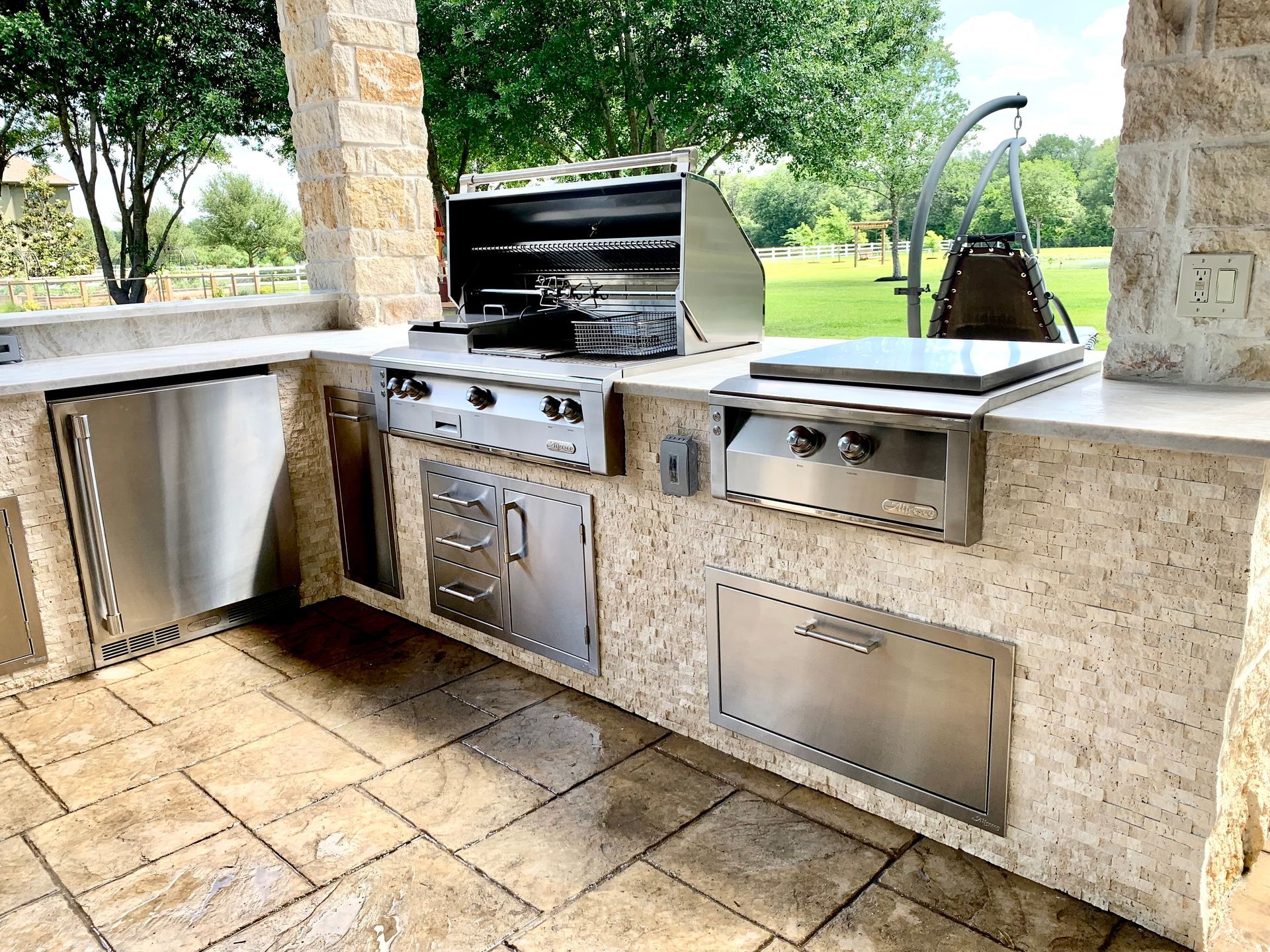 Outdoor stone barbecue kitchen with built-in grill, sink, and storage cabinets under a covered patio