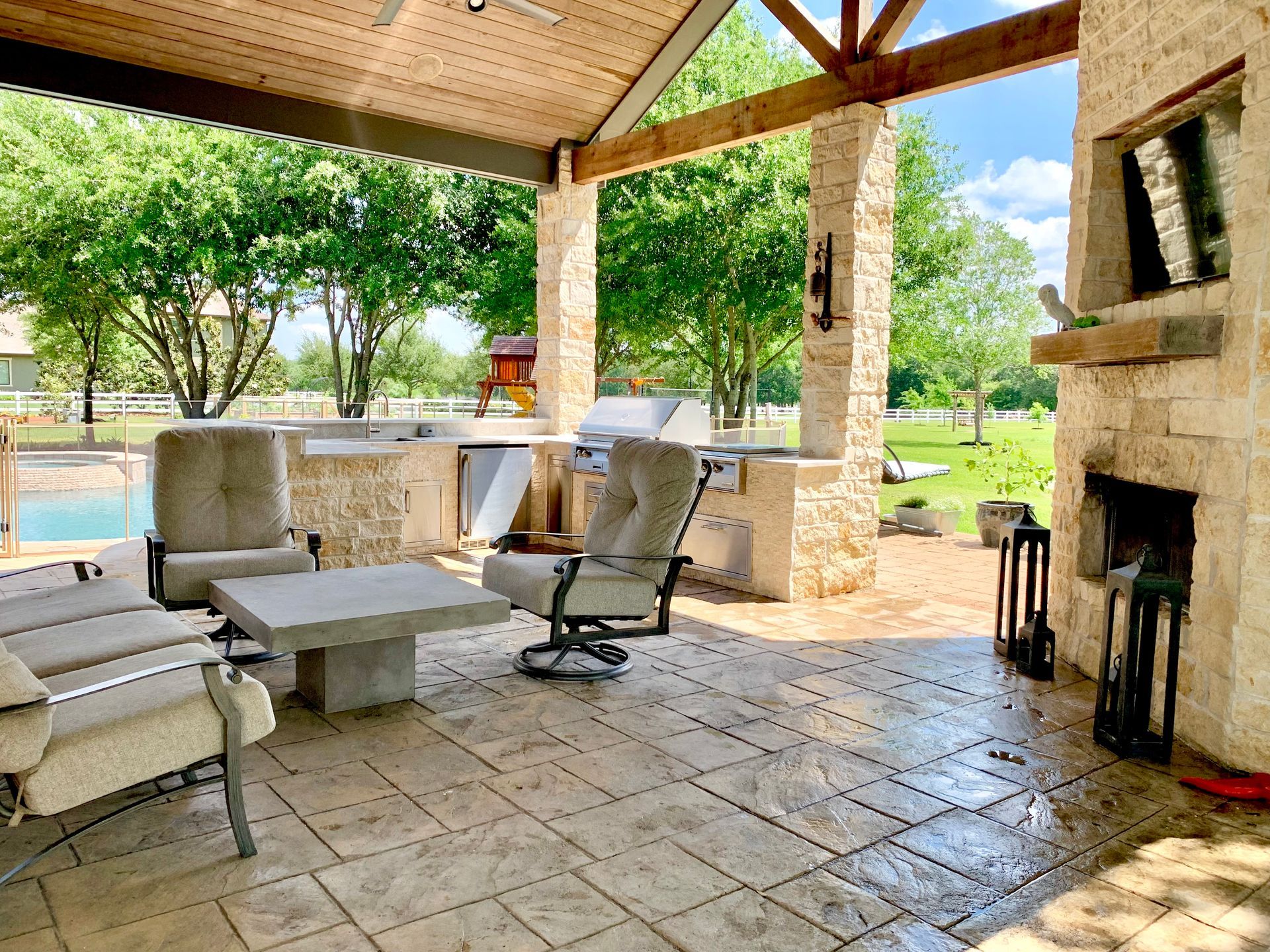 Covered patio with stone columns, wicker seating, and a fireplace overlooking a green lawn.