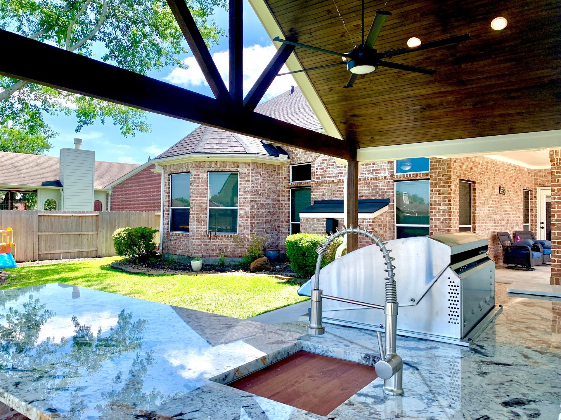 Backyard patio with pergola, outdoor dining table, brick house, and fenced lawn in bright sunlight