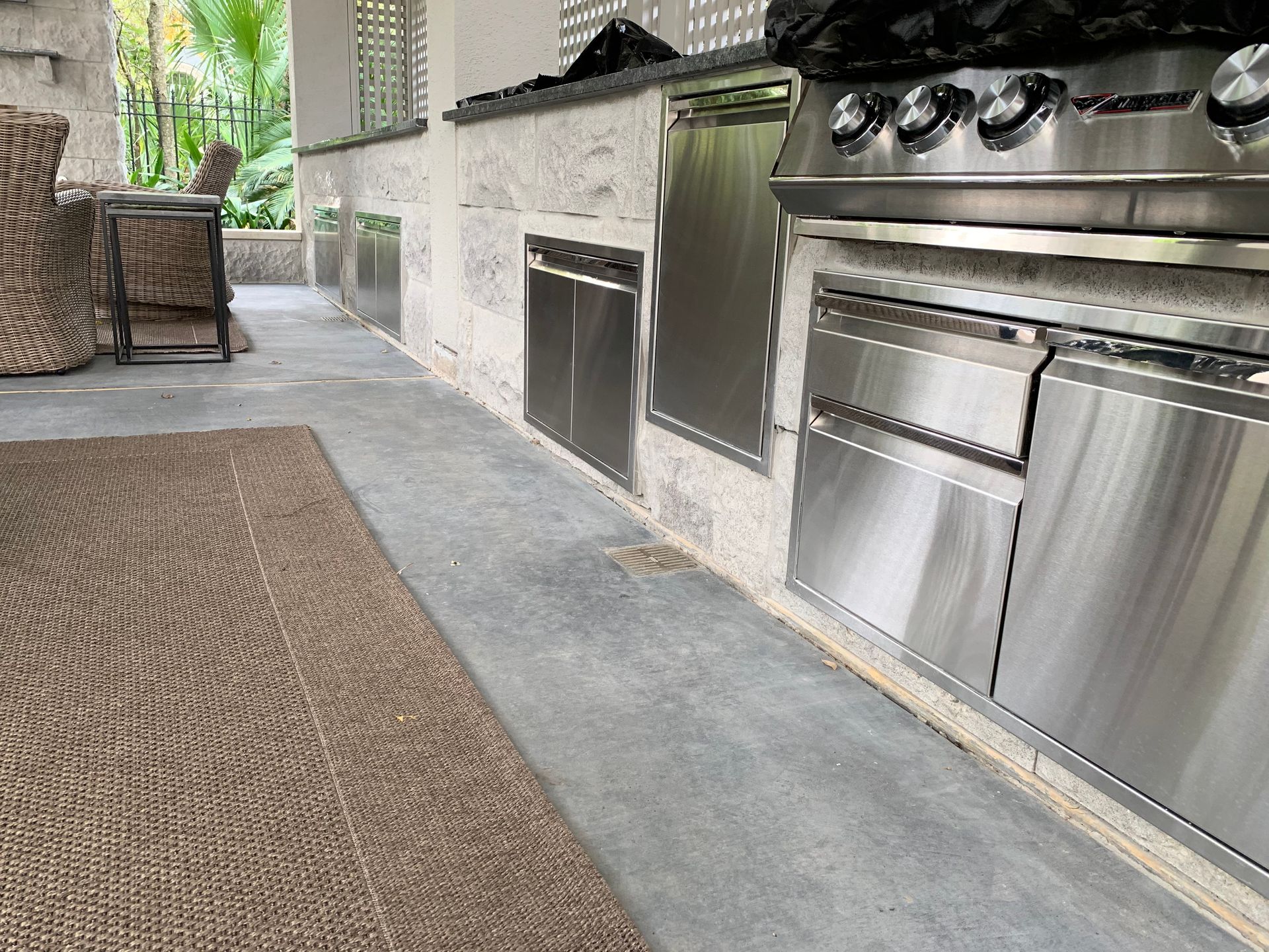 Outdoor patio with a stainless steel grill, stone floor, and woven rug beside chairs and a wall