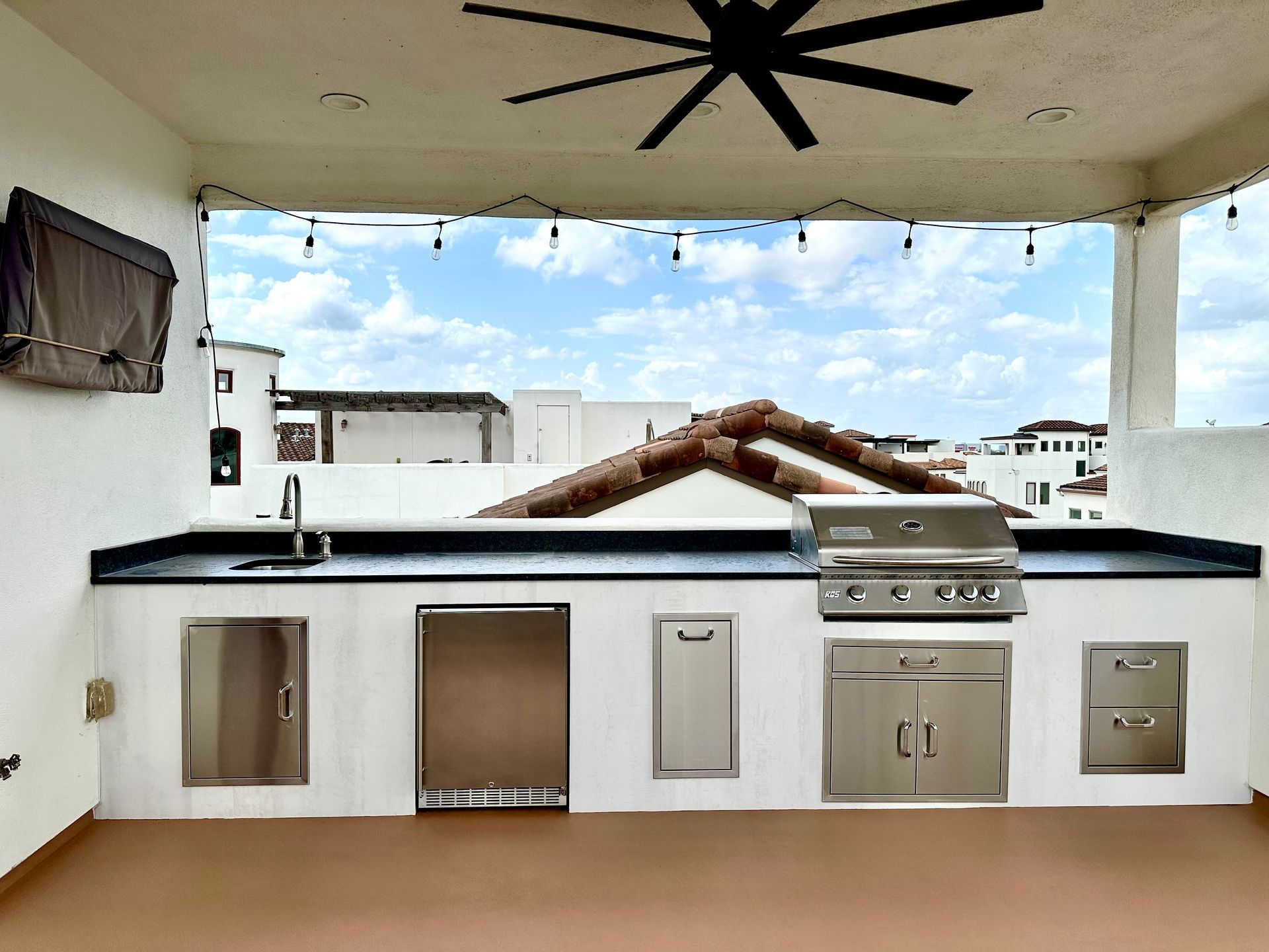 Outdoor rooftop kitchen with grill, sink, and white cabinets under a covered patio.