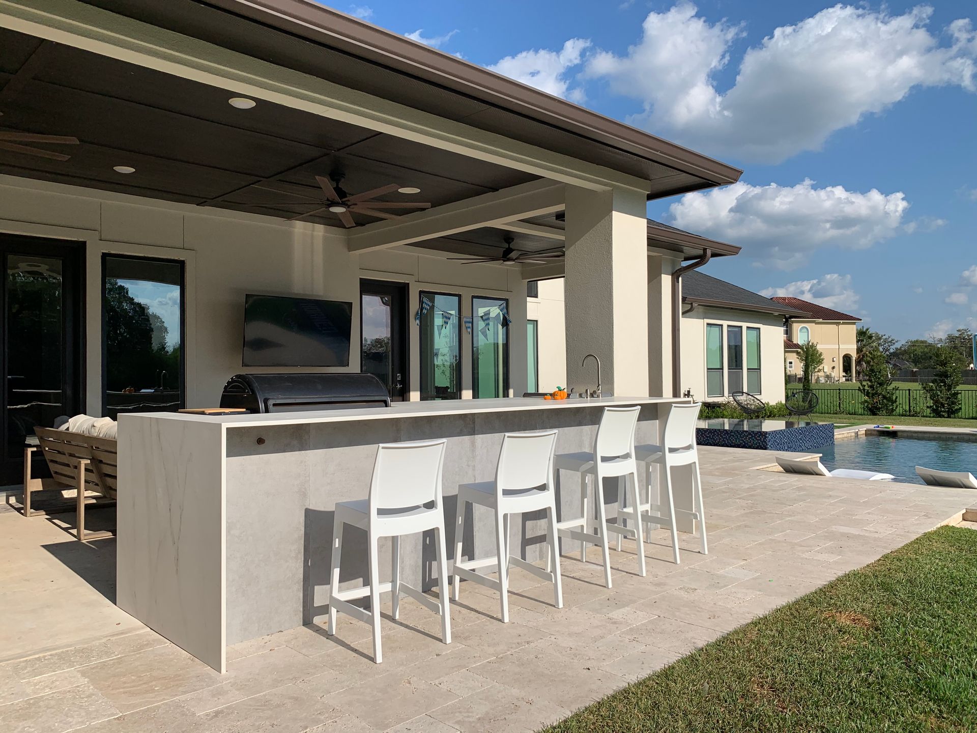 Outdoor patio with white bar stools beside a covered poolside counter and blue swimming pool