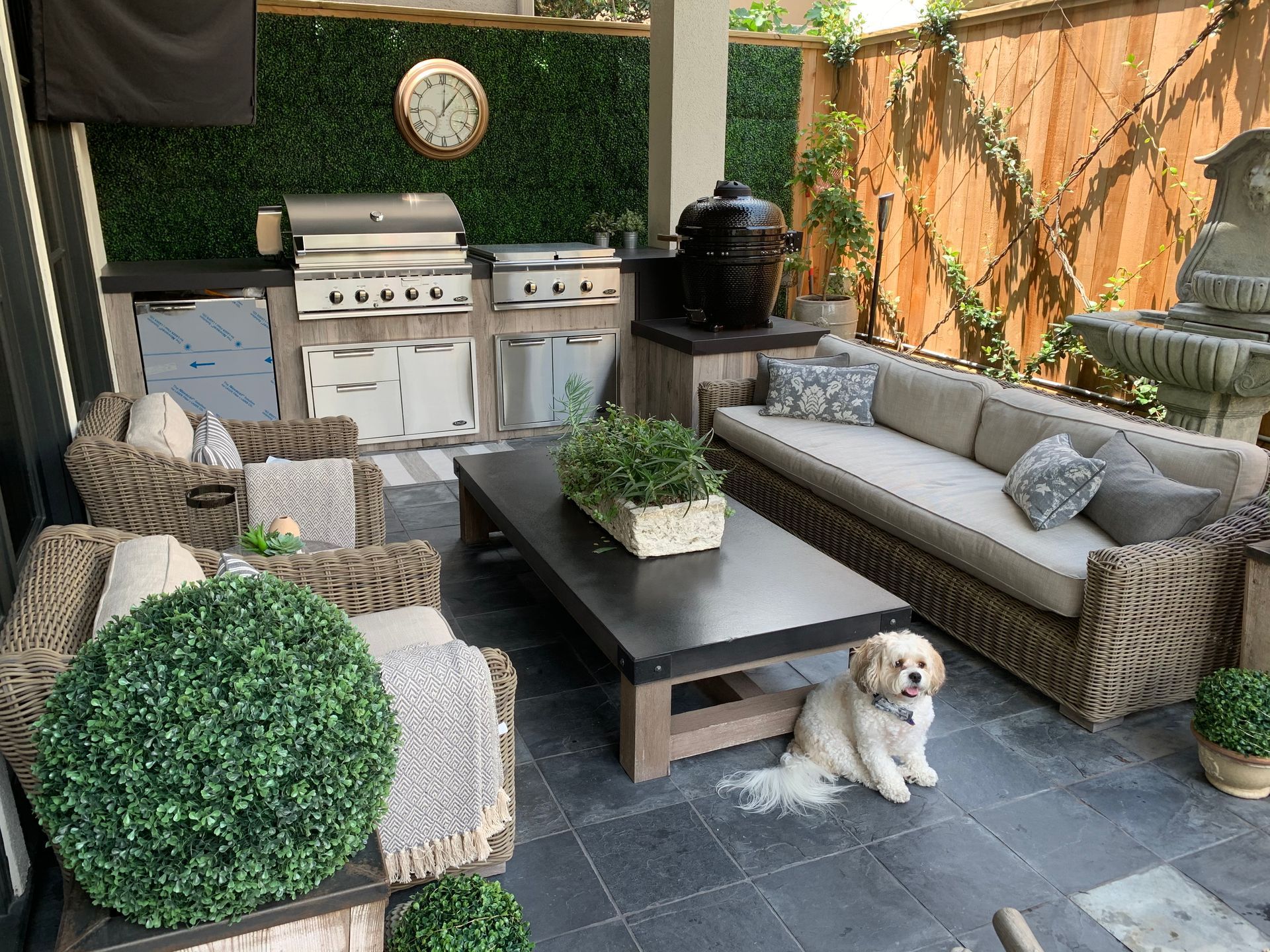 Outdoor patio with wicker sofa, grill, coffee table, potted plants, and a small dog on dark tile floor