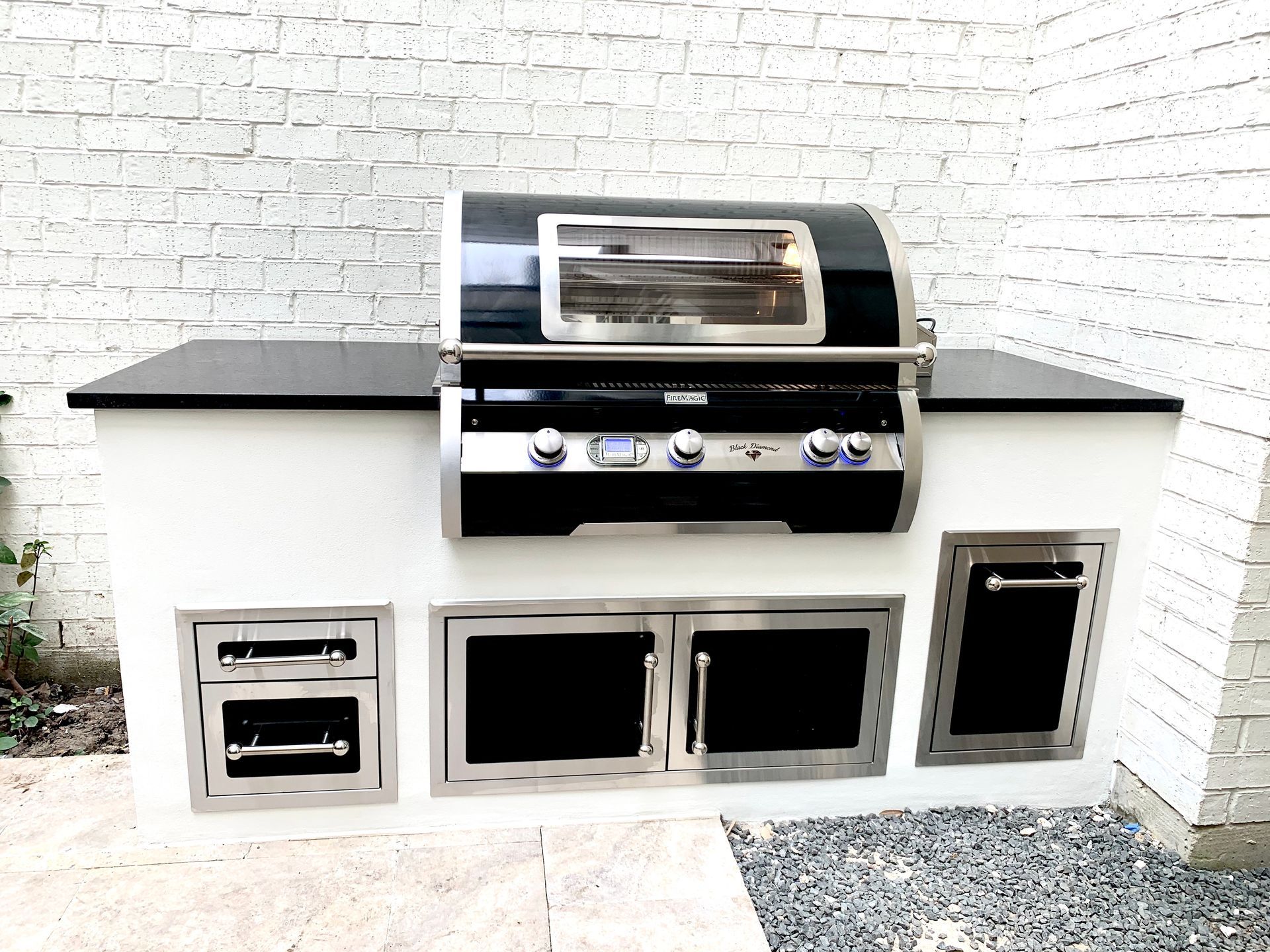 Outdoor built-in stainless steel grill island with side cabinets against a white brick wall.
