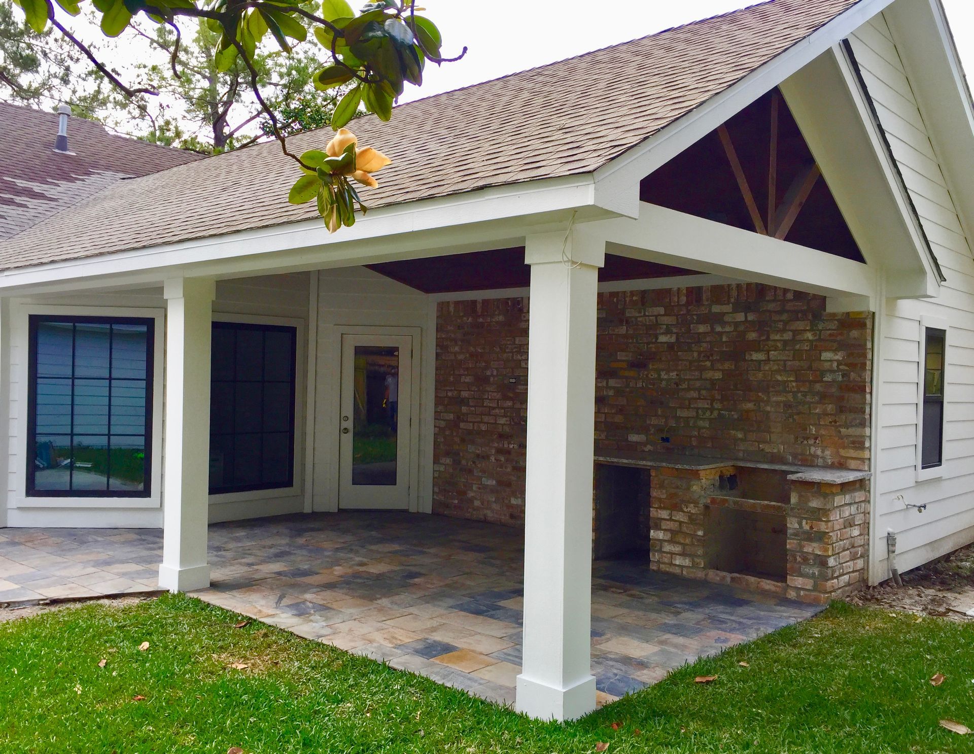 Covered patio with white columns, brick walls, and stone flooring beside a house with a lawn.