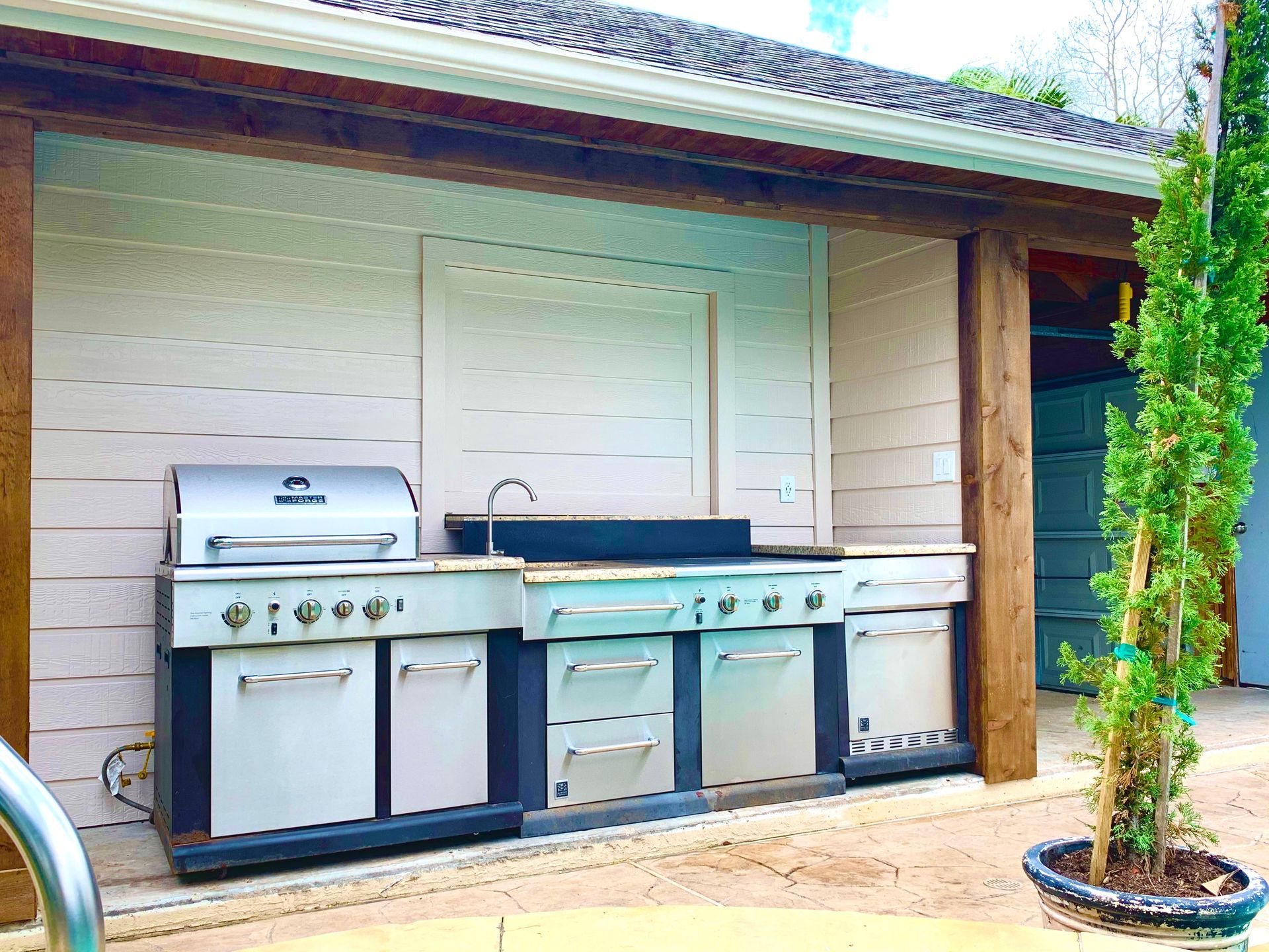 Outdoor grill and sink station beside a house, with a silver barbecue and blue cabinets under a roof.
