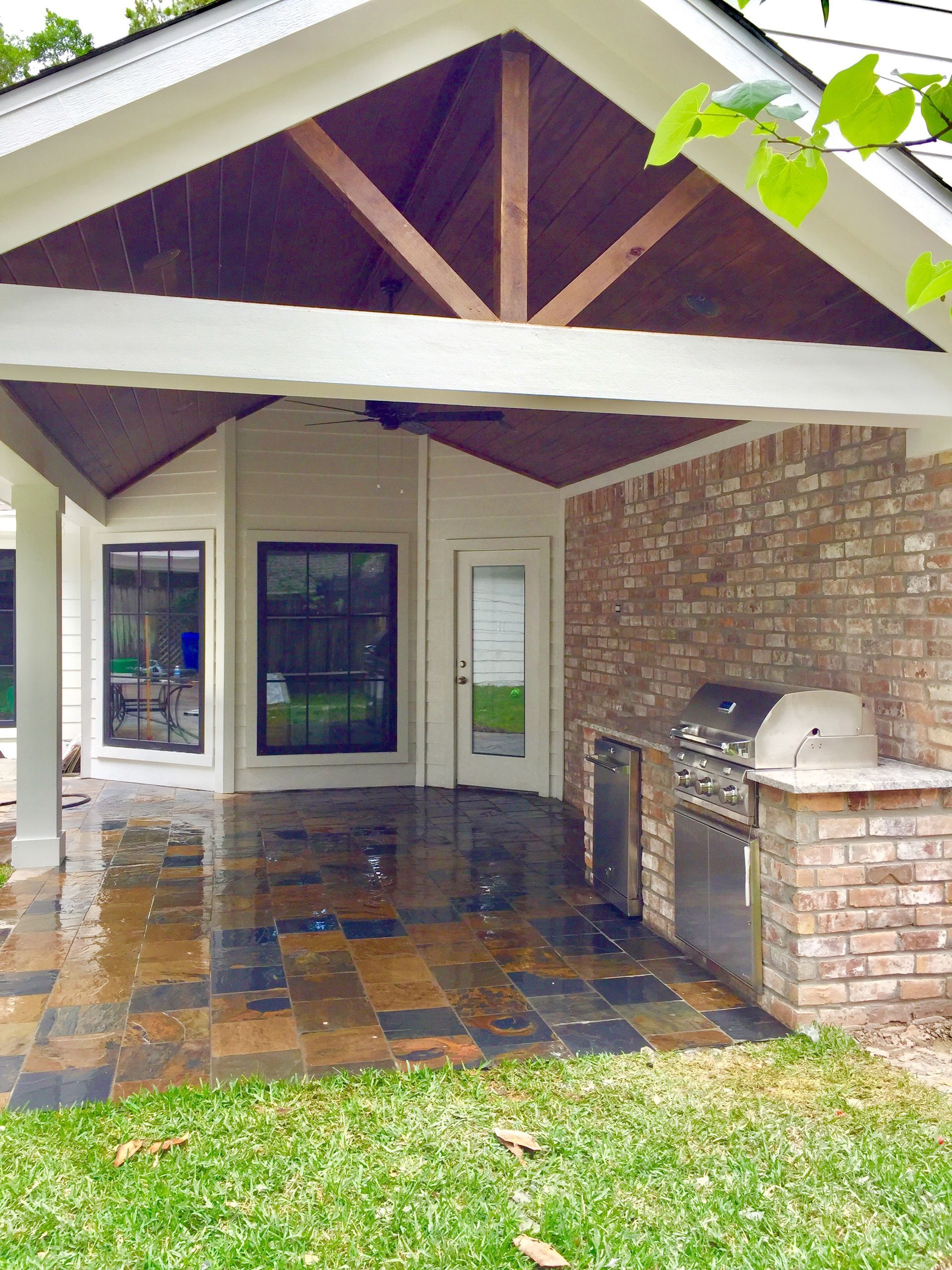 Covered brick patio with wet tile floor, wooden gabled roof, and built-in grill beside glass doors.