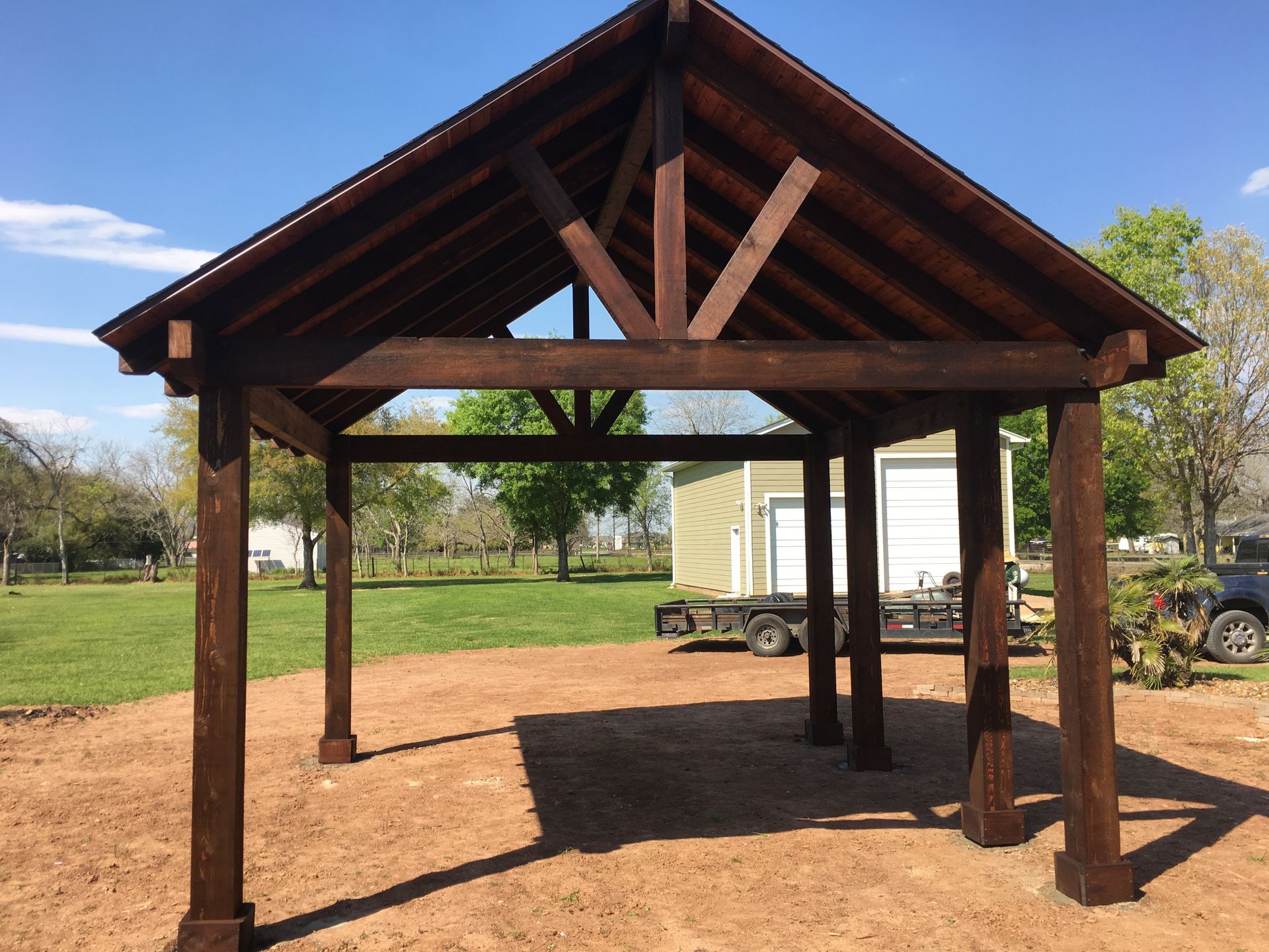Open-sided wooden pavilion with a peaked roof on a gravel lot in a grassy park