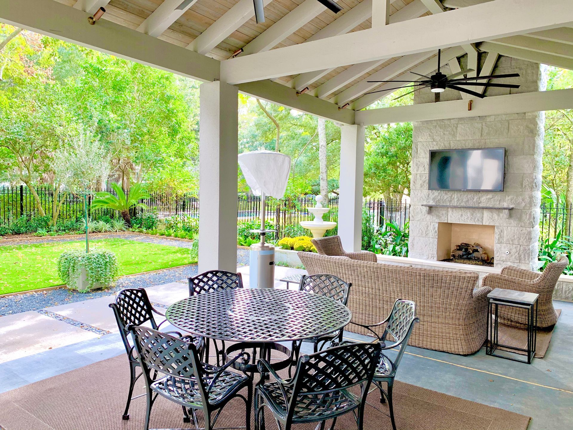 Covered patio with dining table, chairs, and outdoor seating beside a stone fireplace and garden view.
