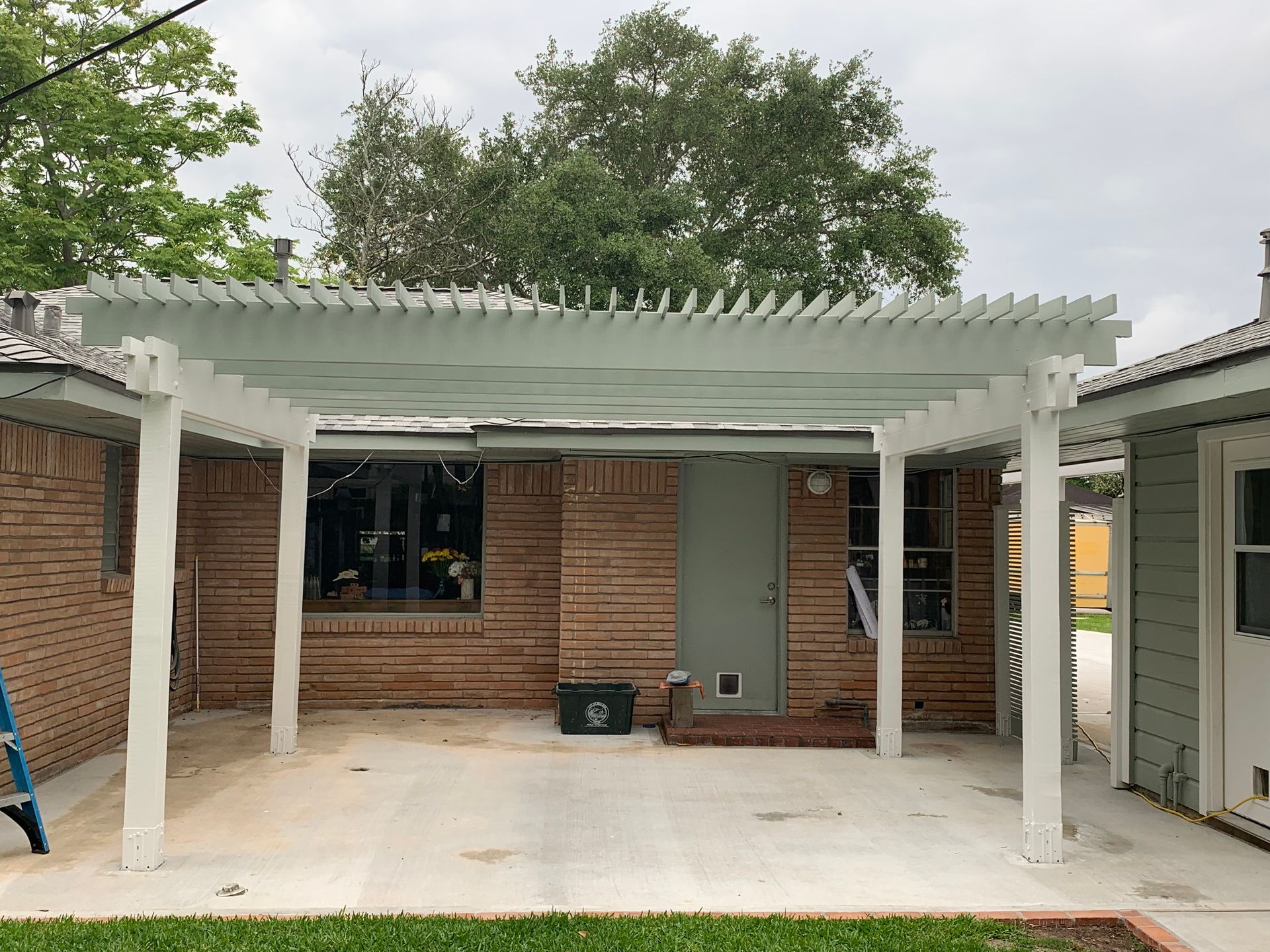 Covered brick patio with white pergola posts, concrete floor, and doors on both sides under cloudy sky