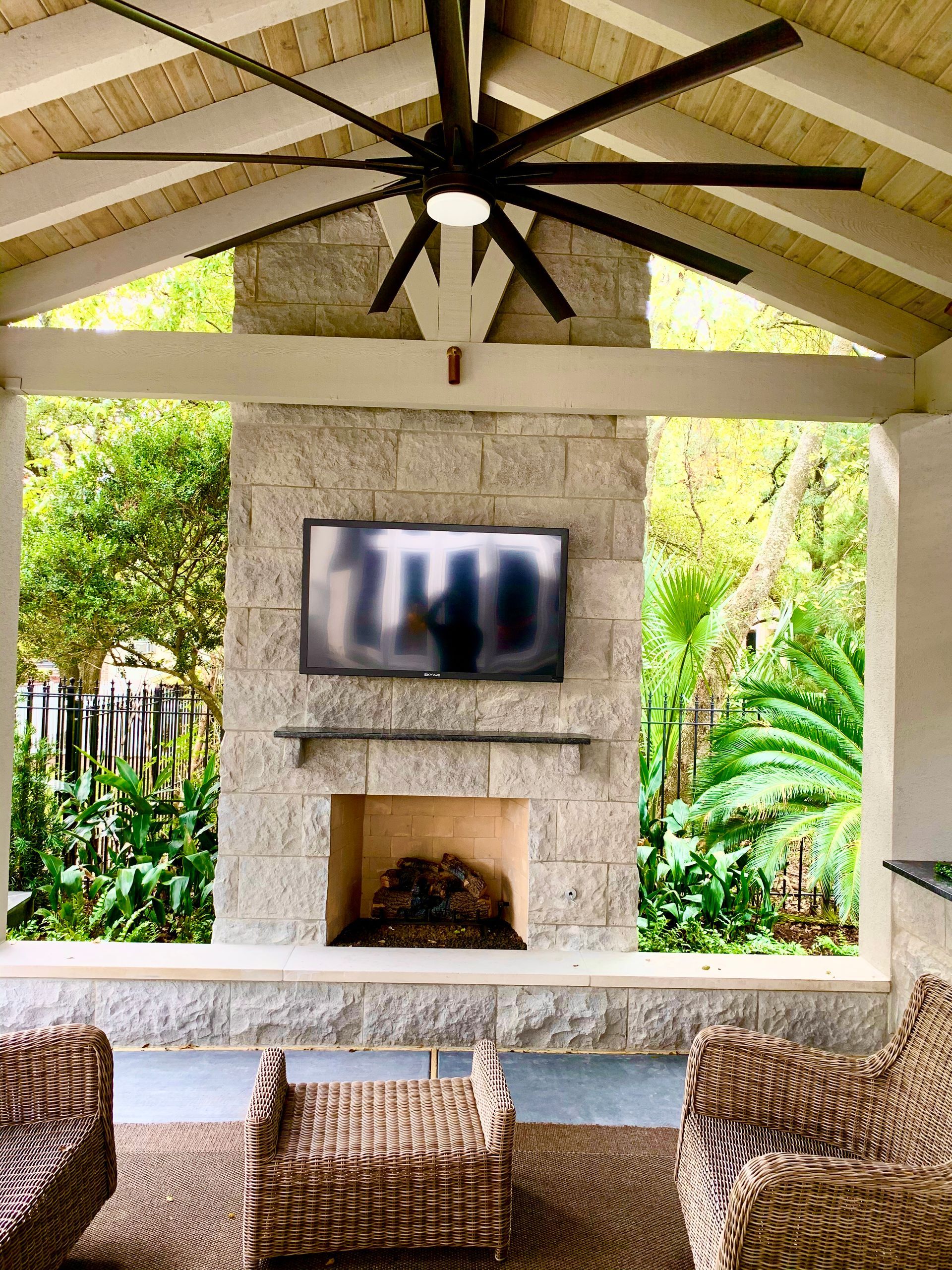 Covered patio with stone fireplace, wall-mounted TV, wicker chairs, and ceiling fan overlooking greenery